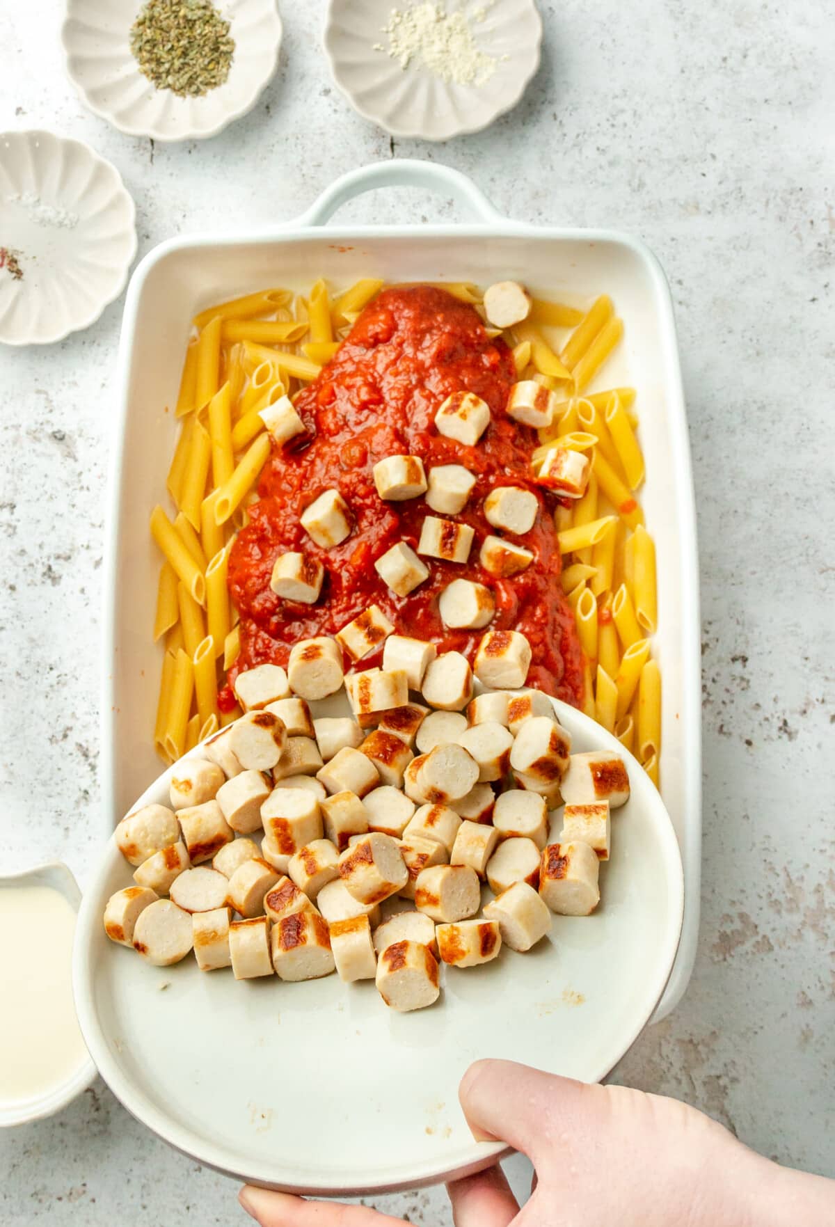 Sliced chicken sausage is tossed over marinara sauce and penne pasta in a baking dish on a light grey colored surface.