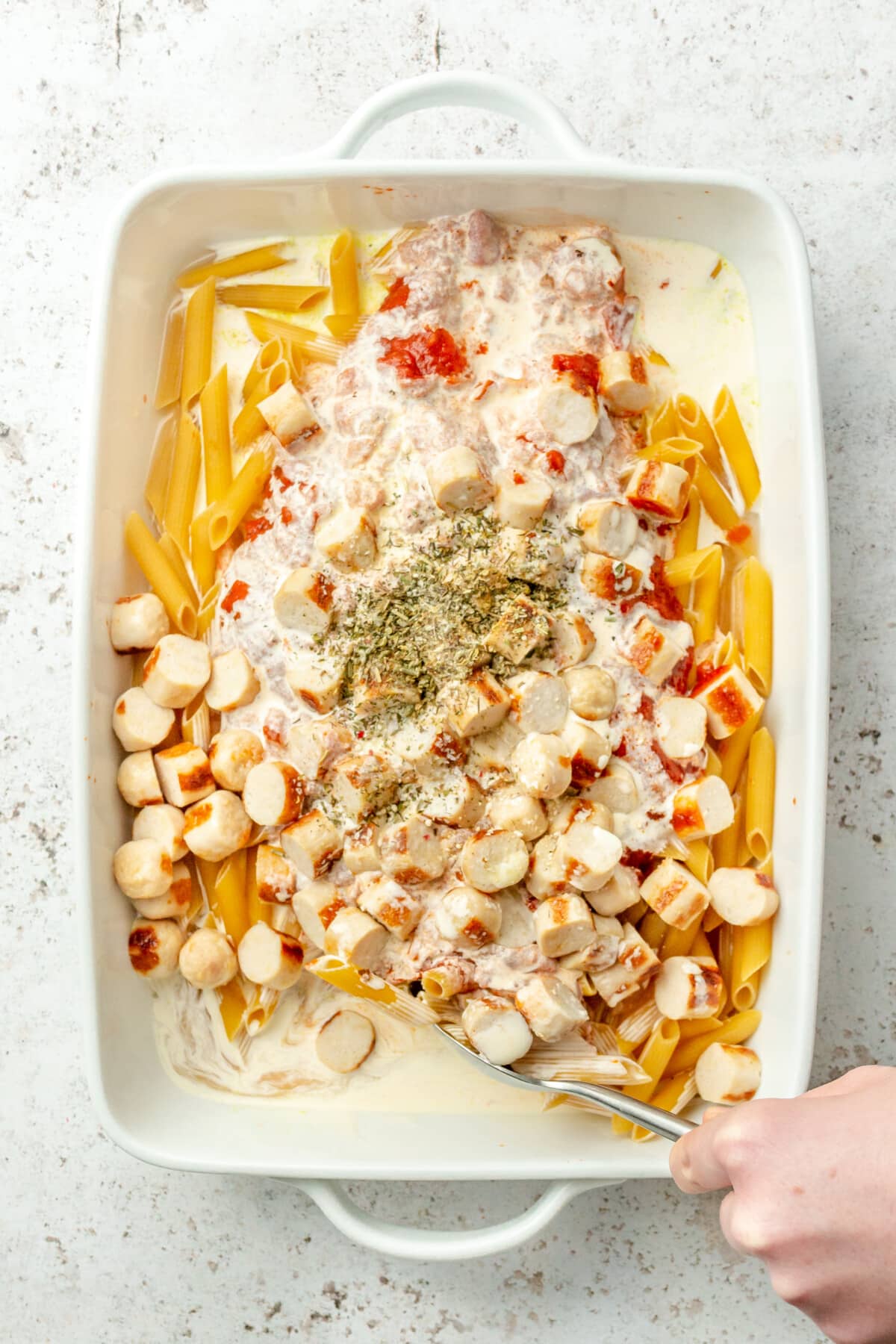 No bake pasta bake ingredients are stirred together in a baking dish on a light grey colored surface.