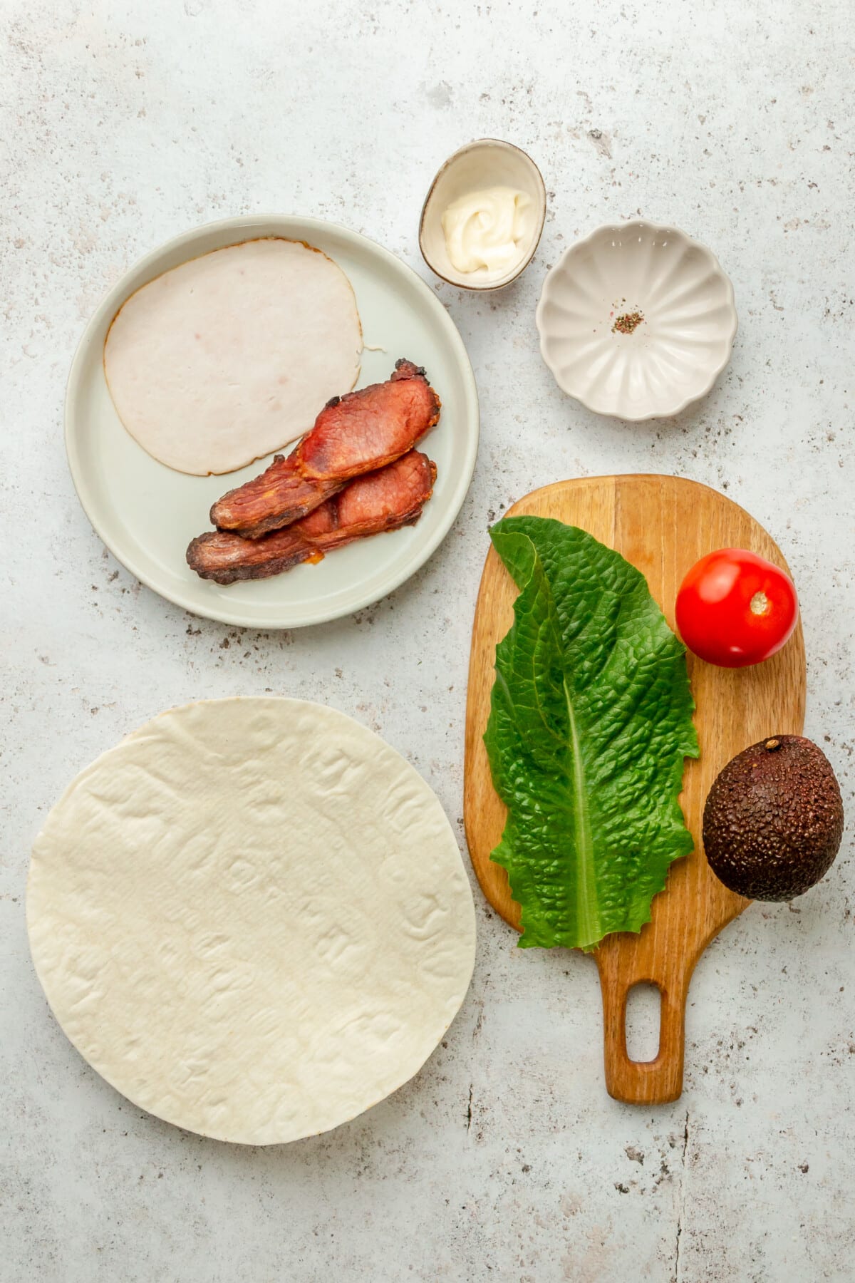 Ingredients for turkey avocado wraps sit in a variety of bowls and plates on a light grey surface.