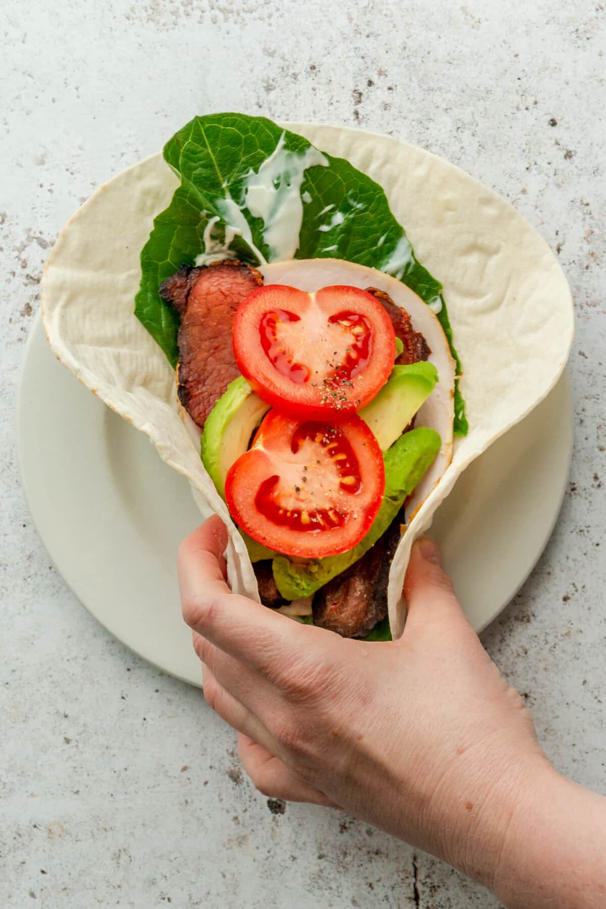 A turkey avocado wrap is picked up from a white ceramic plate on a light grey surface.