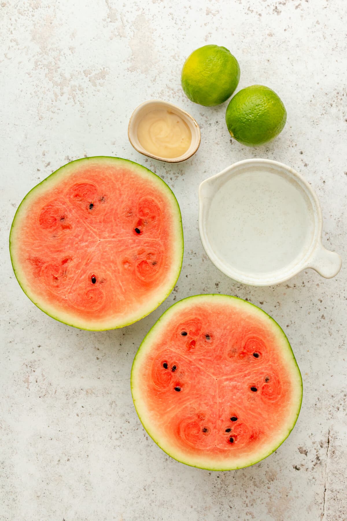 Ingredients for frozen watermelon maragaritas sit in a variety of bowls on a light grey surface.
