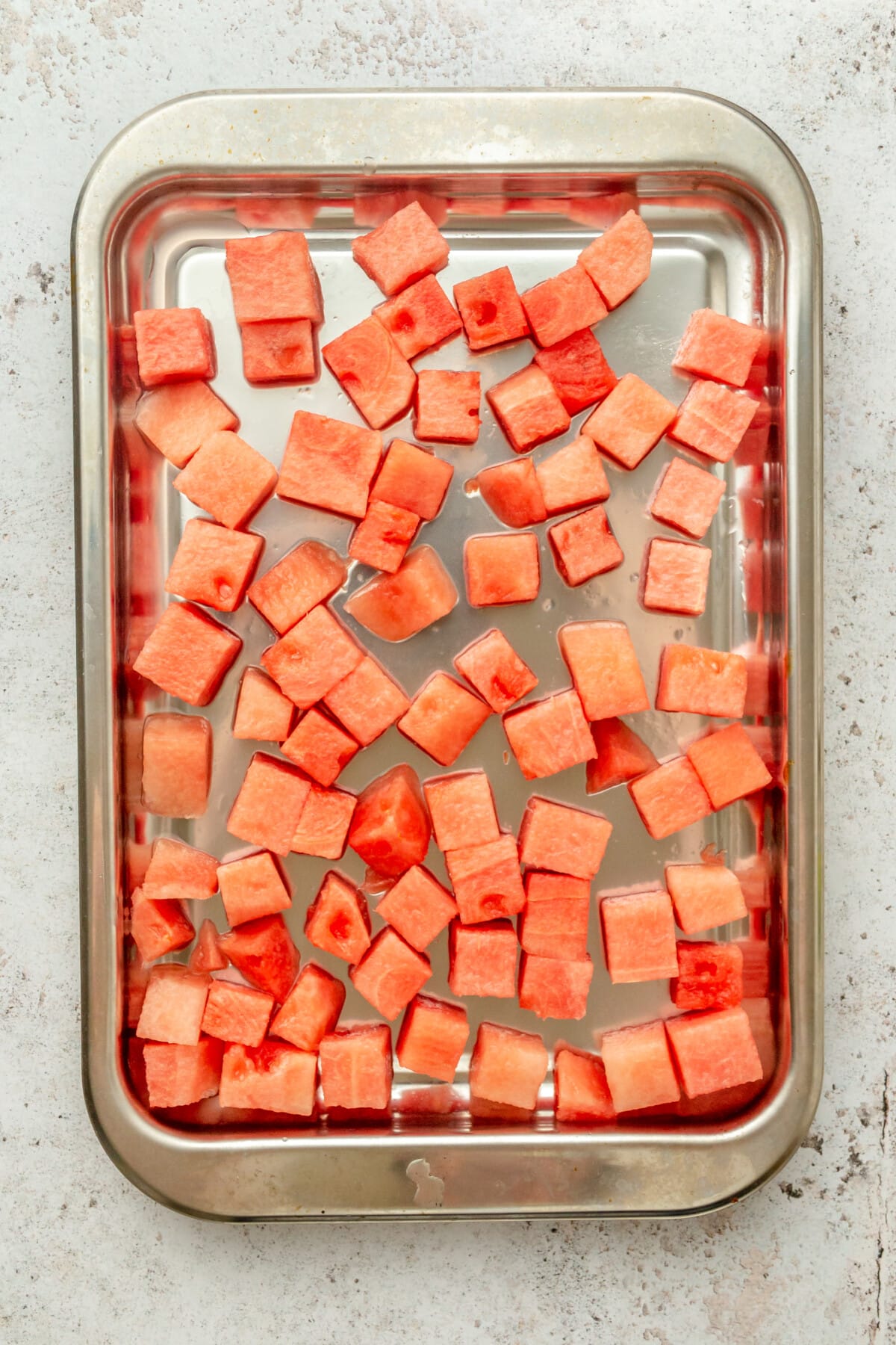 Cubes of fresh watermelon sit on a tray to be frozen on a light grey surface.