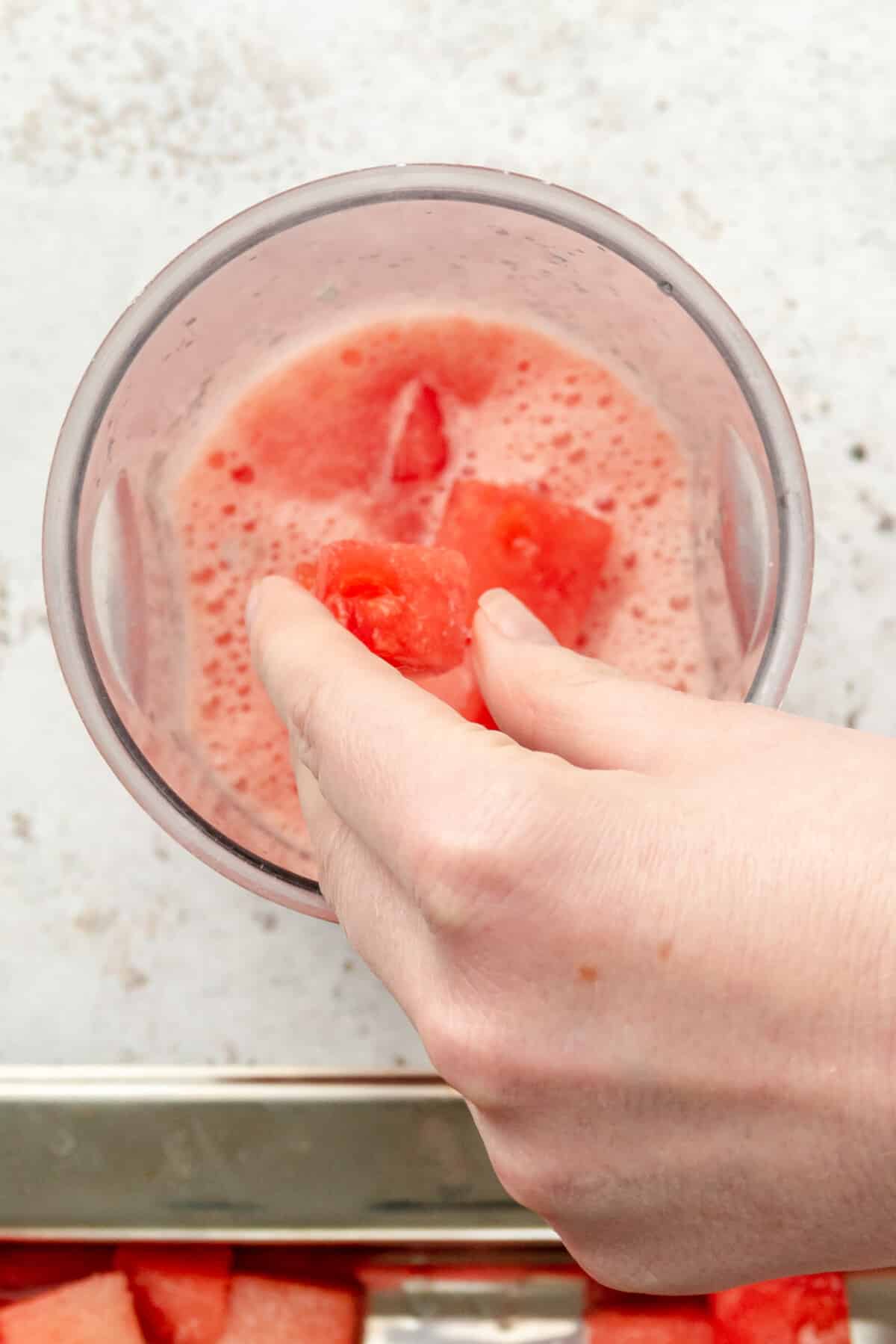 Cubes of frozen margaritas are tossed into a blender beaker for a watermelon margaritas on a light grey surface.