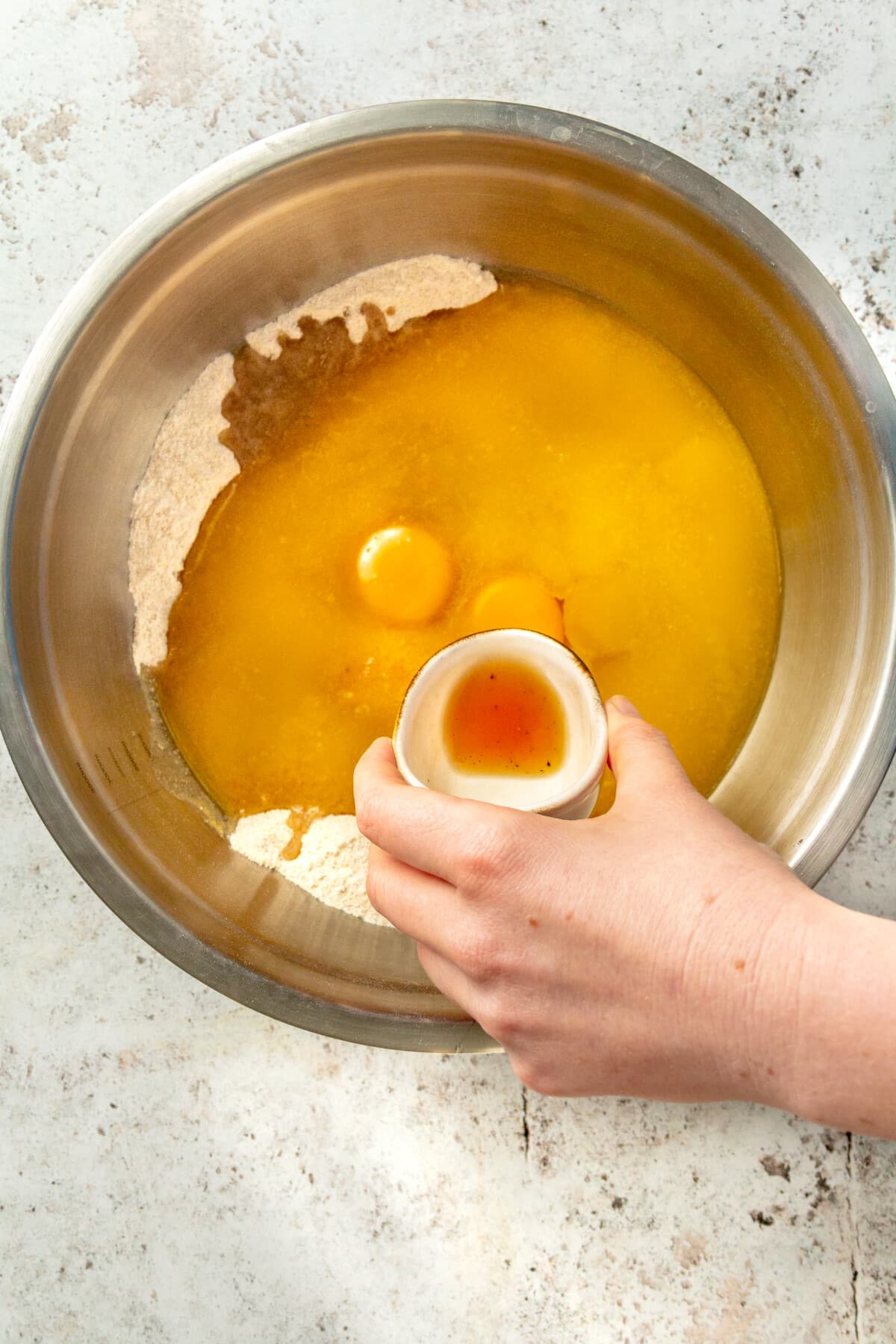 Vanilla extract is shown being added to a large metal mixing bowl which holds eggs and several other ingredients.