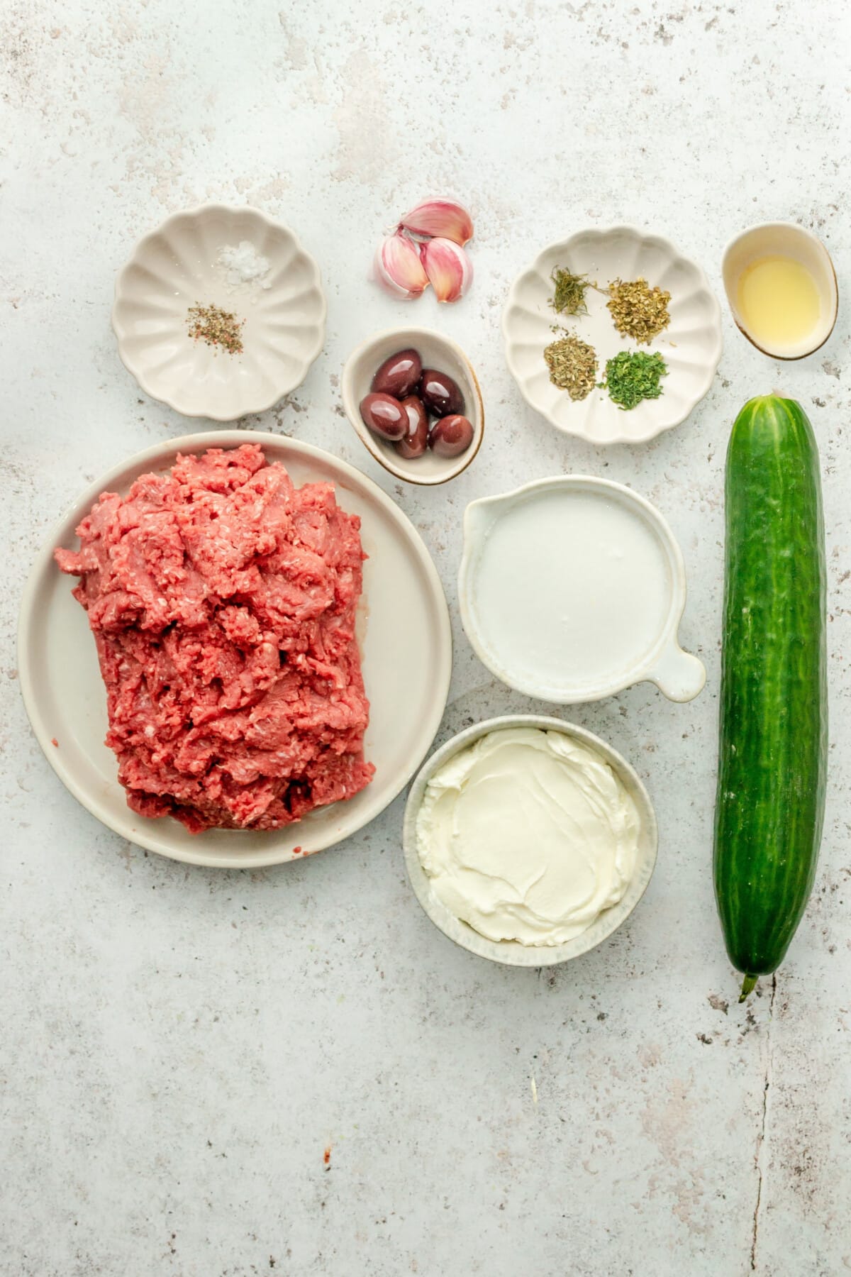 Ingredients for Greek inspired meatballs with tzatziki sit in a variety of bowls and plates on a light grey surface.