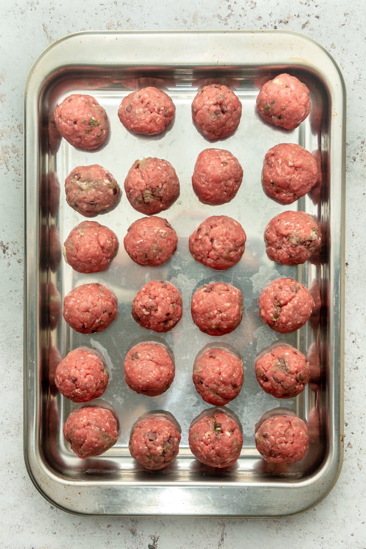 Unbaked Greek inspired meatballs sit on a rimmed baking stainless steel tray on a light grey surface.