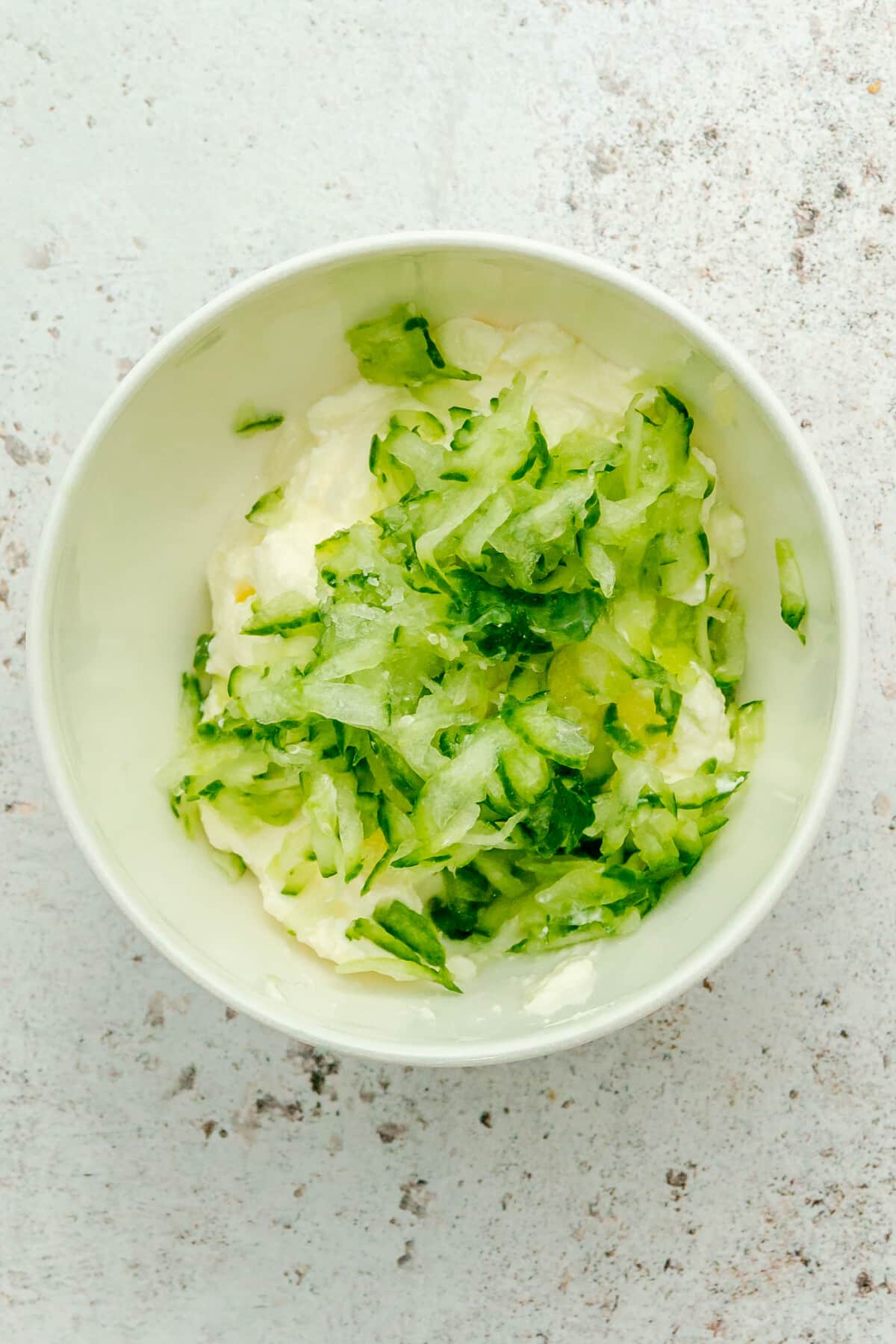 Grated cucumber sits on top of yogurt in a white ceramic bowl Unbaked Greek inspired meatballs sit on a light grey surface.