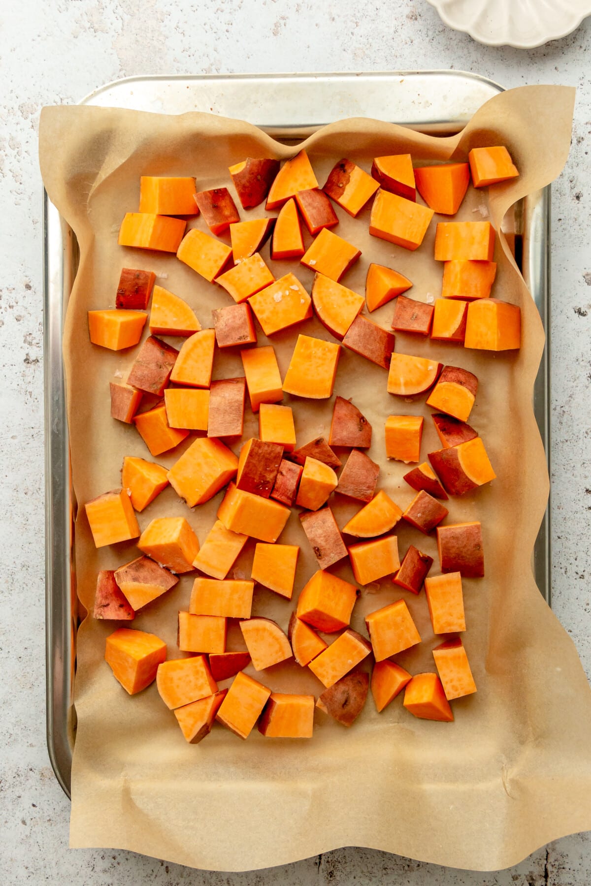 Cubes of sweet potato sit on a lined and rimmed stainless steel baking sheet on a light grey surface.