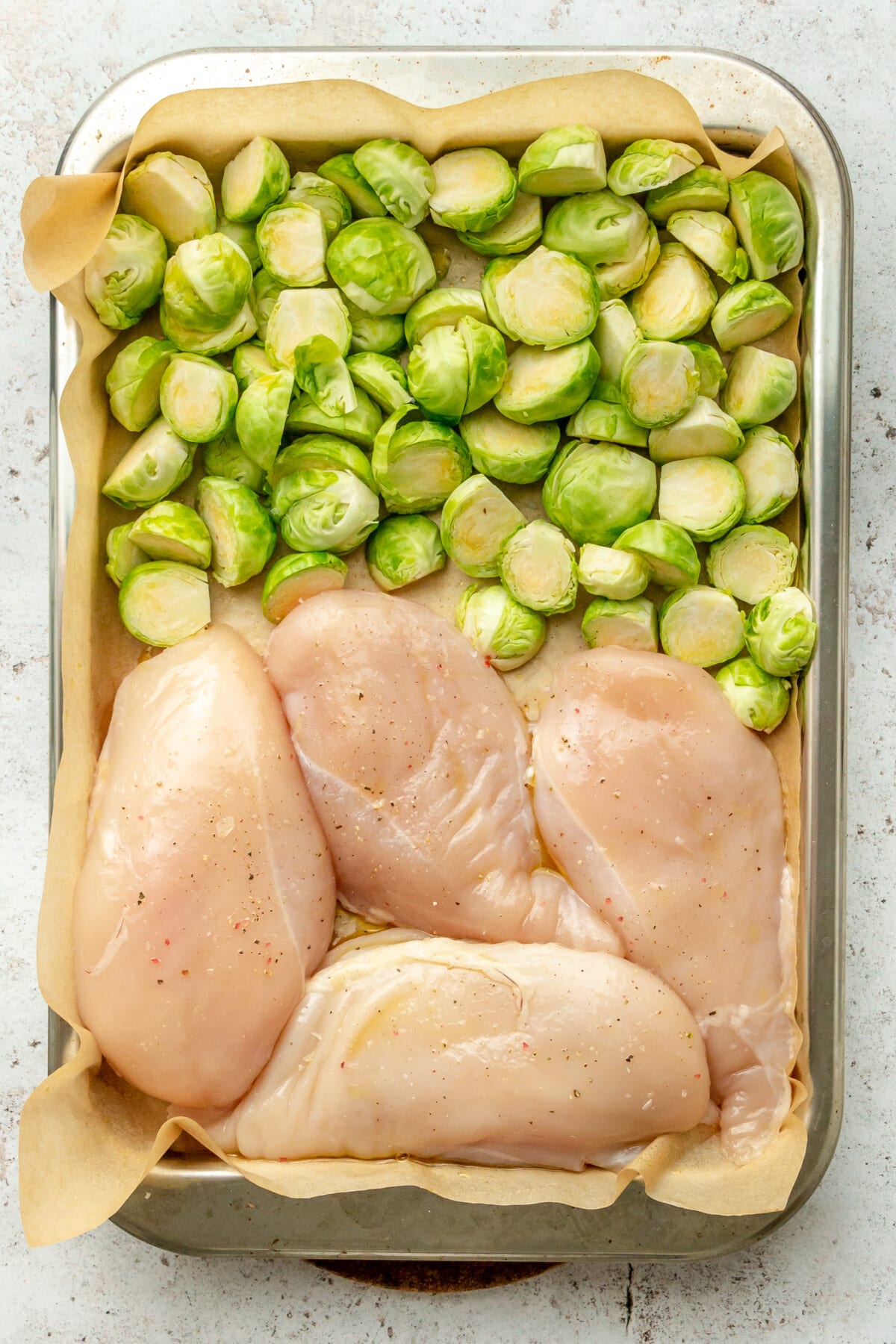 Chicken breastes and halved brussel sprouts sit on a rimmed lined baking sheet on a light grey surface.