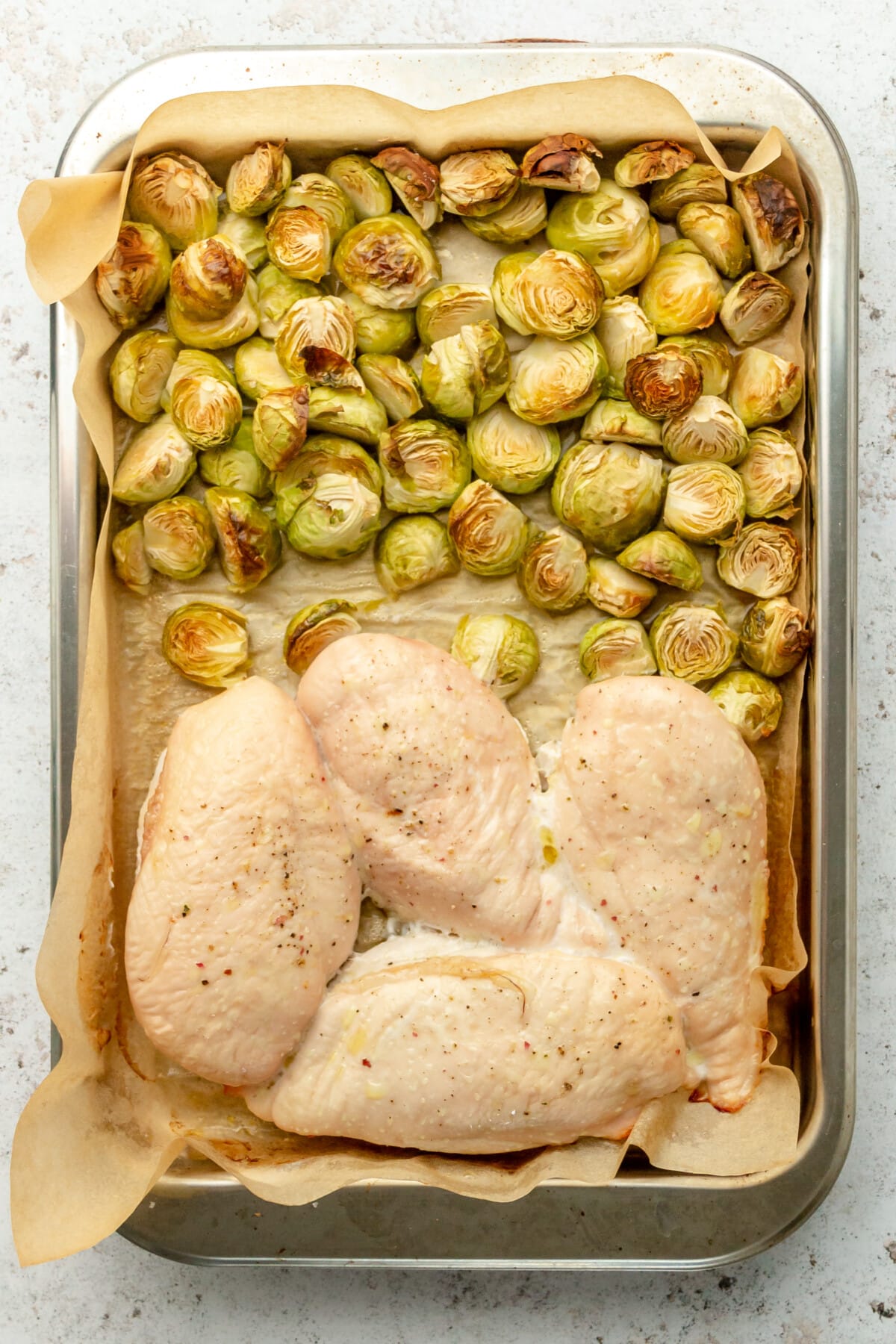 Baked chicken breasts and brussel sprouts sit on a lined rimmed baking sheet on a light grey surface.