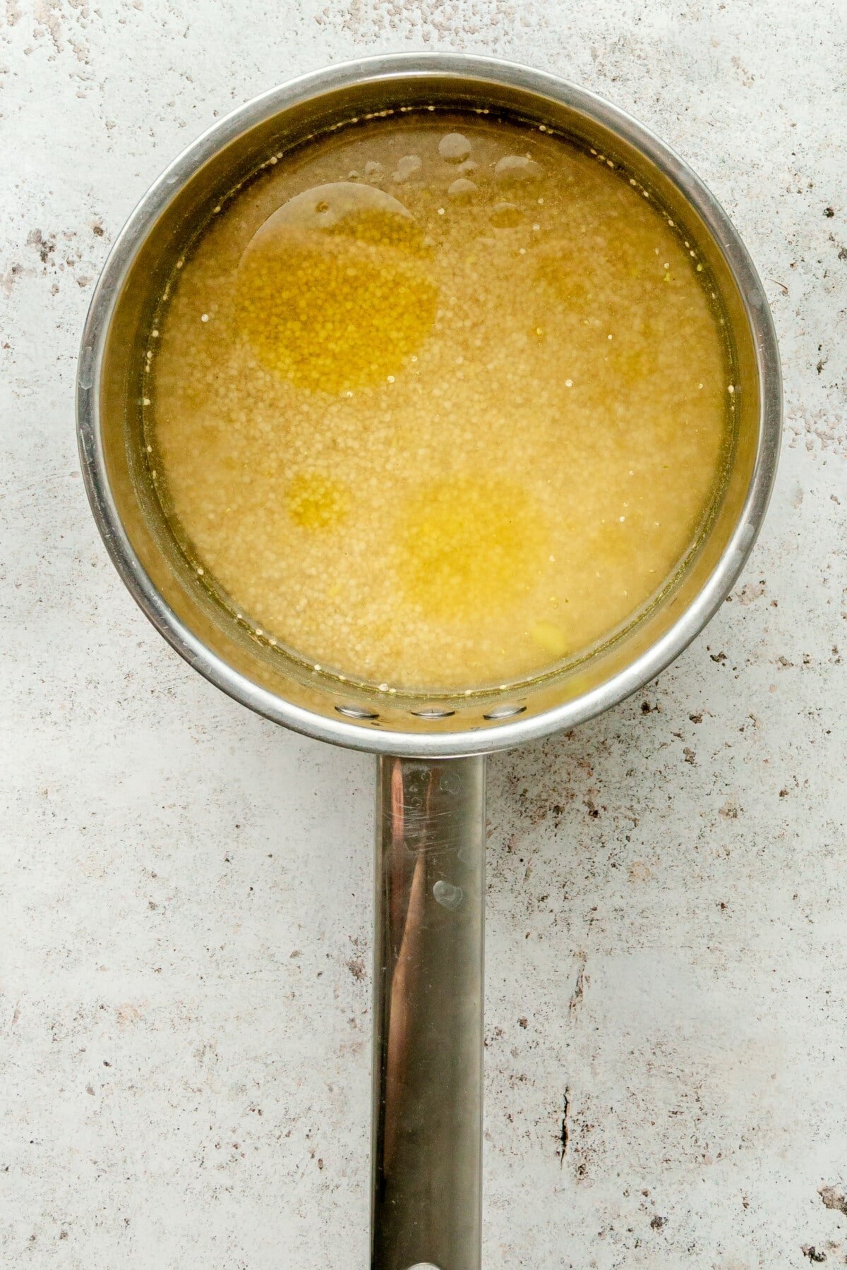 Quinoa sits in a small saucepan covered in broth on a light grey surface.