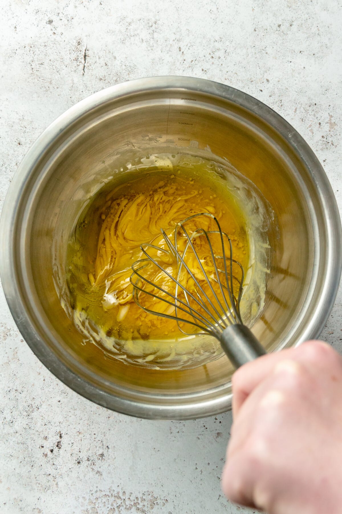 A honey mustard sauce is whisked in a stainless steel bowl on a light grey colored surface.