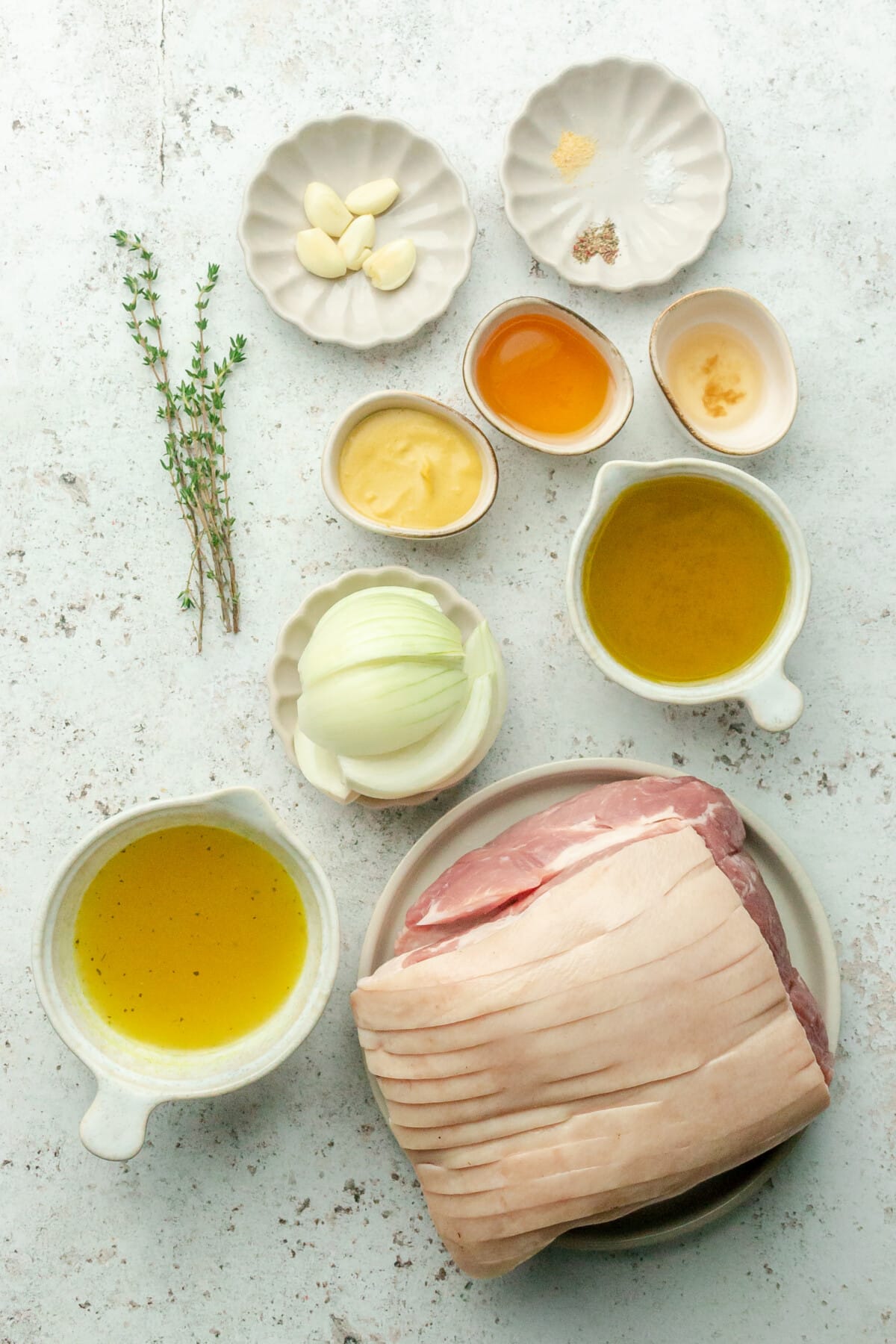 Ingredients for honey mustard pork loin roast sit in a variety of bowls and plates on a light grey surface.