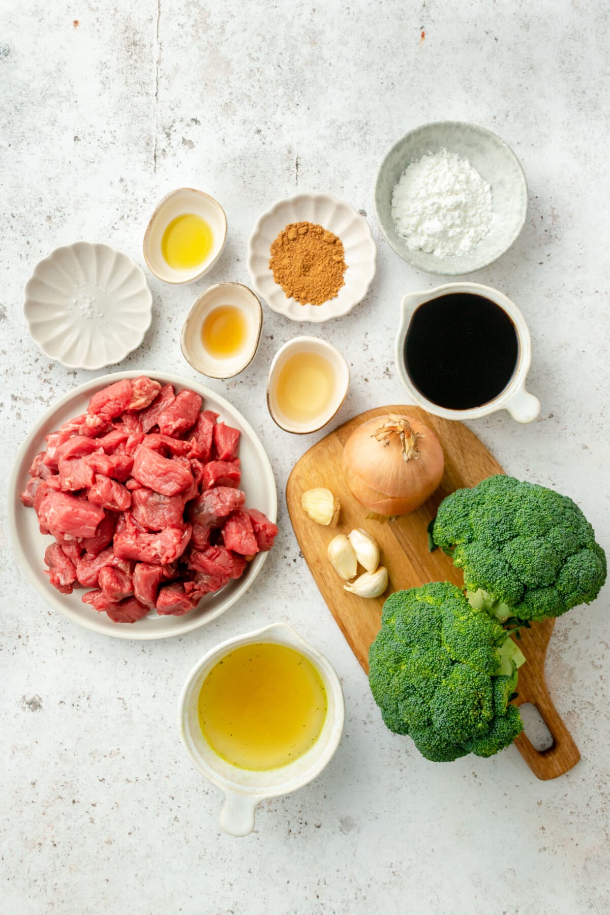 Ingredients for Instant Pot Beef Brocoli are laid out in a variety of bowls and plates on a light grey colored surface.