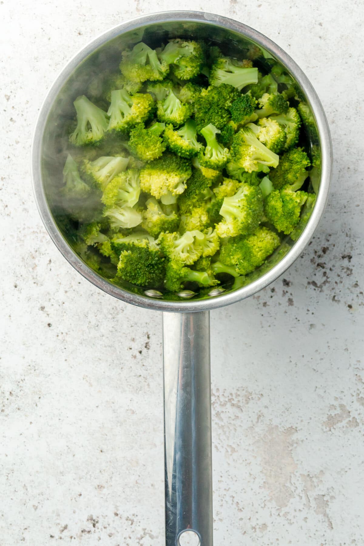 Cooked broccoli florets sit in a saucepan with a little steam coming off on a light grey colored surface.