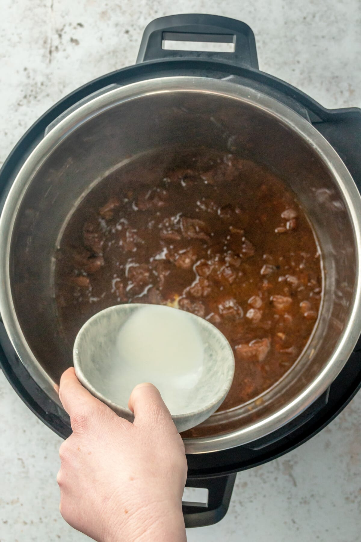 A corn starch slurry is added to a beef dish in an instant pot on a light grey colored surface.
