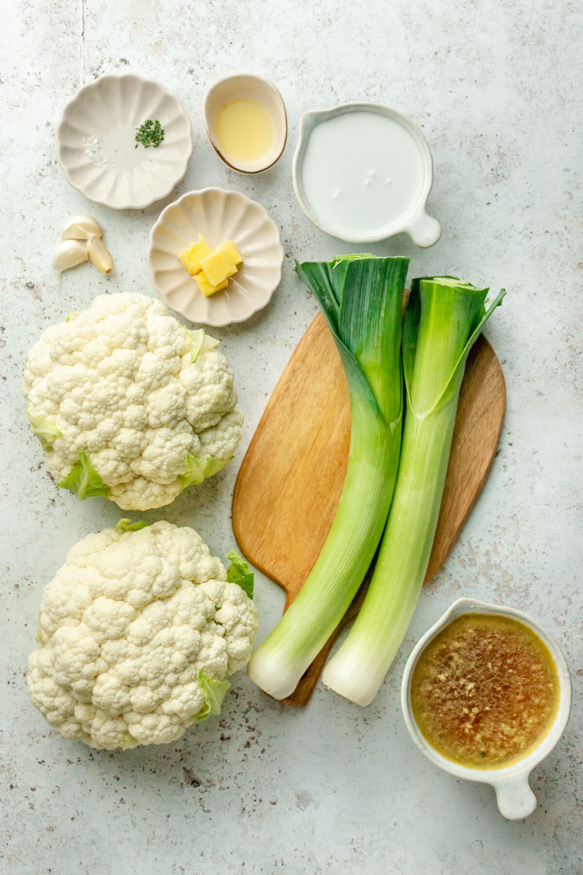 Ingredients for cauliflower soup with leek sit in a variety of bowls and plates on a light grey surface.