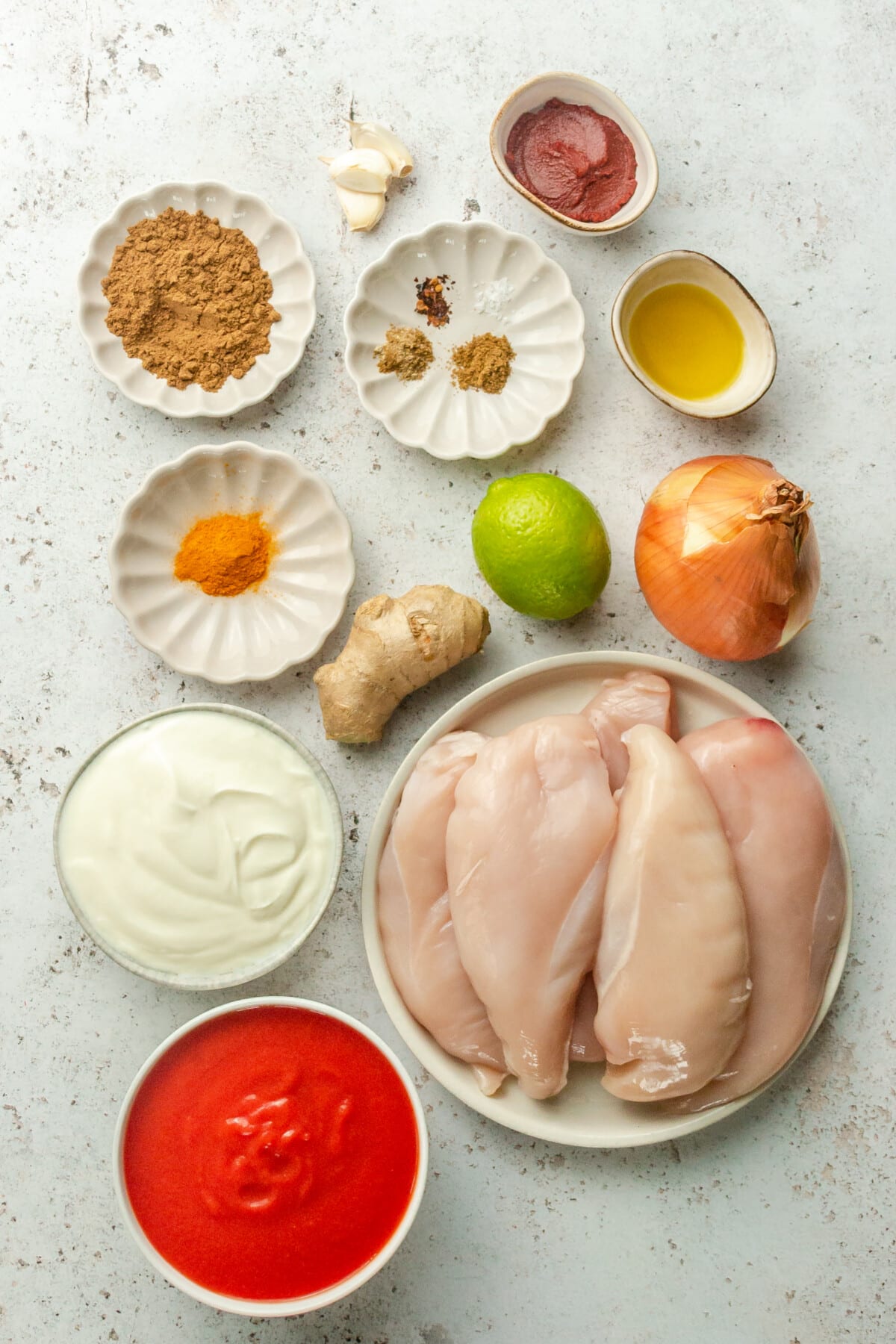 Ingredients for chicken tikka masala sit in a variety of bowls and plates on a light grey surface.