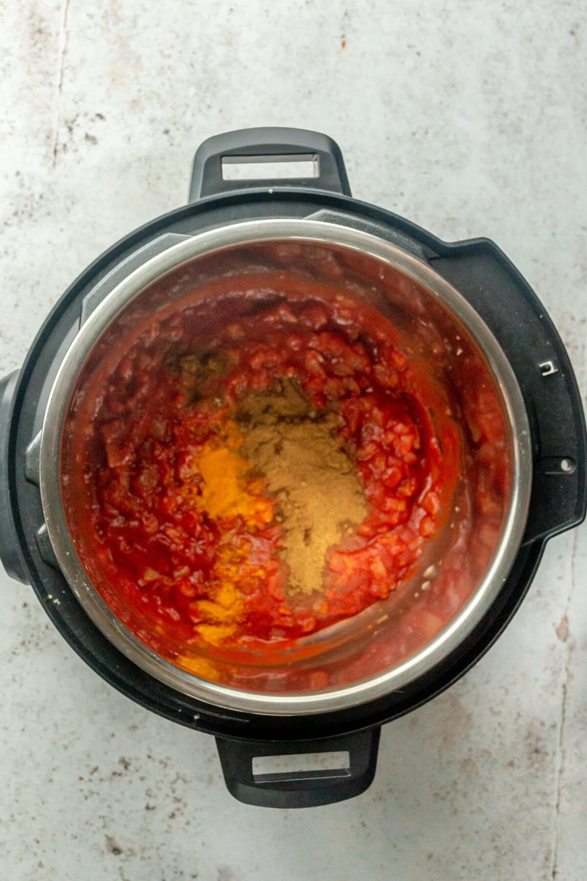 Ground spices sit on top of tomato sauce in an instant pot on a light grey surface.