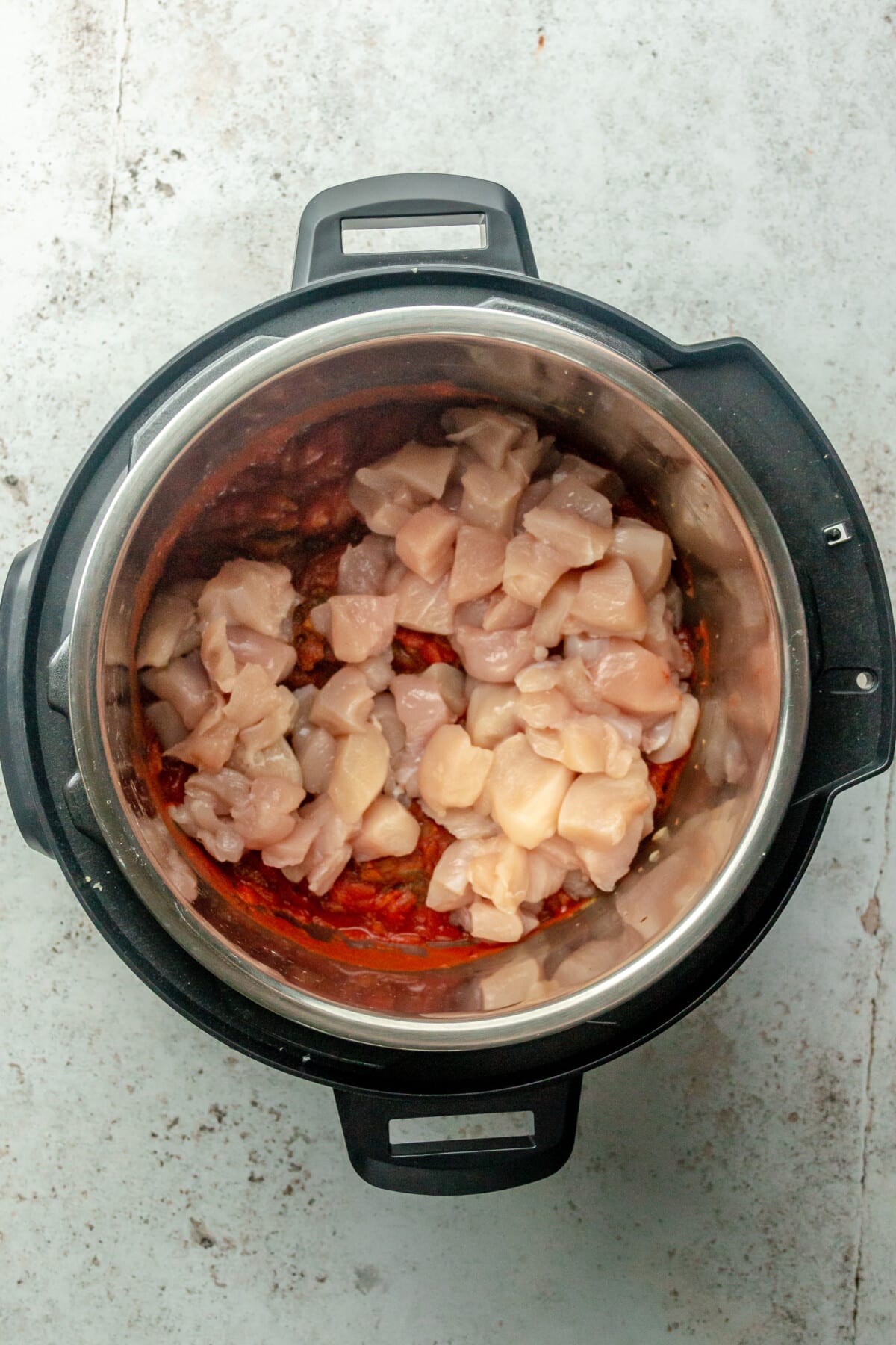 Diced chicken pieces sit on top of tomato sauce in an instant pot on a light grey surface.