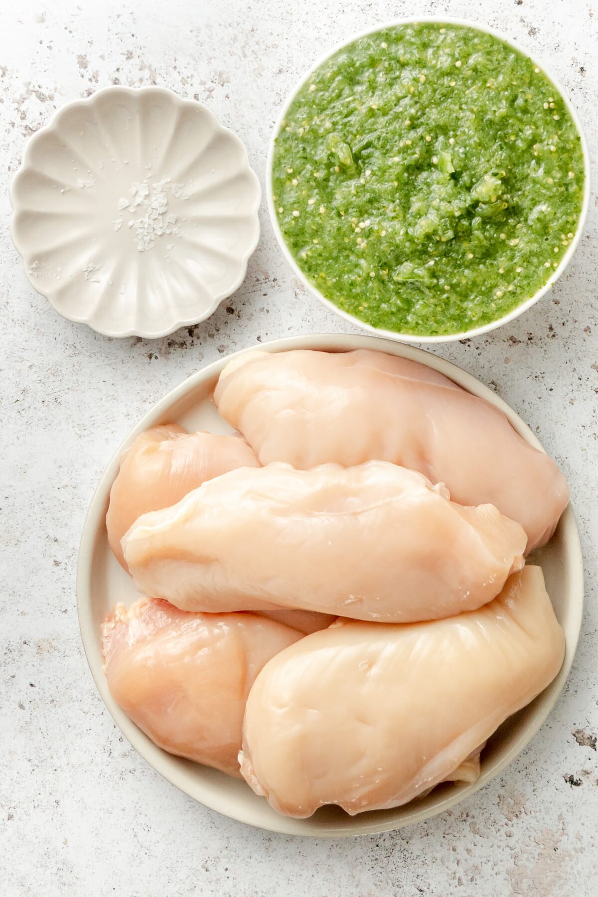 Ingredients for instant pot salsa chicken sit in a variety of plates on a light grey surface.