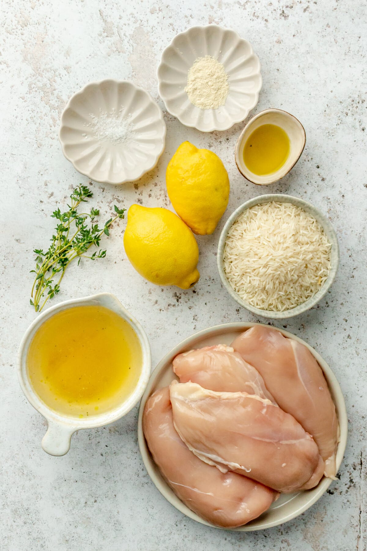 Ingredients for lemon chicken and rice sit in a variety of bowls and plates on a light grey surface.