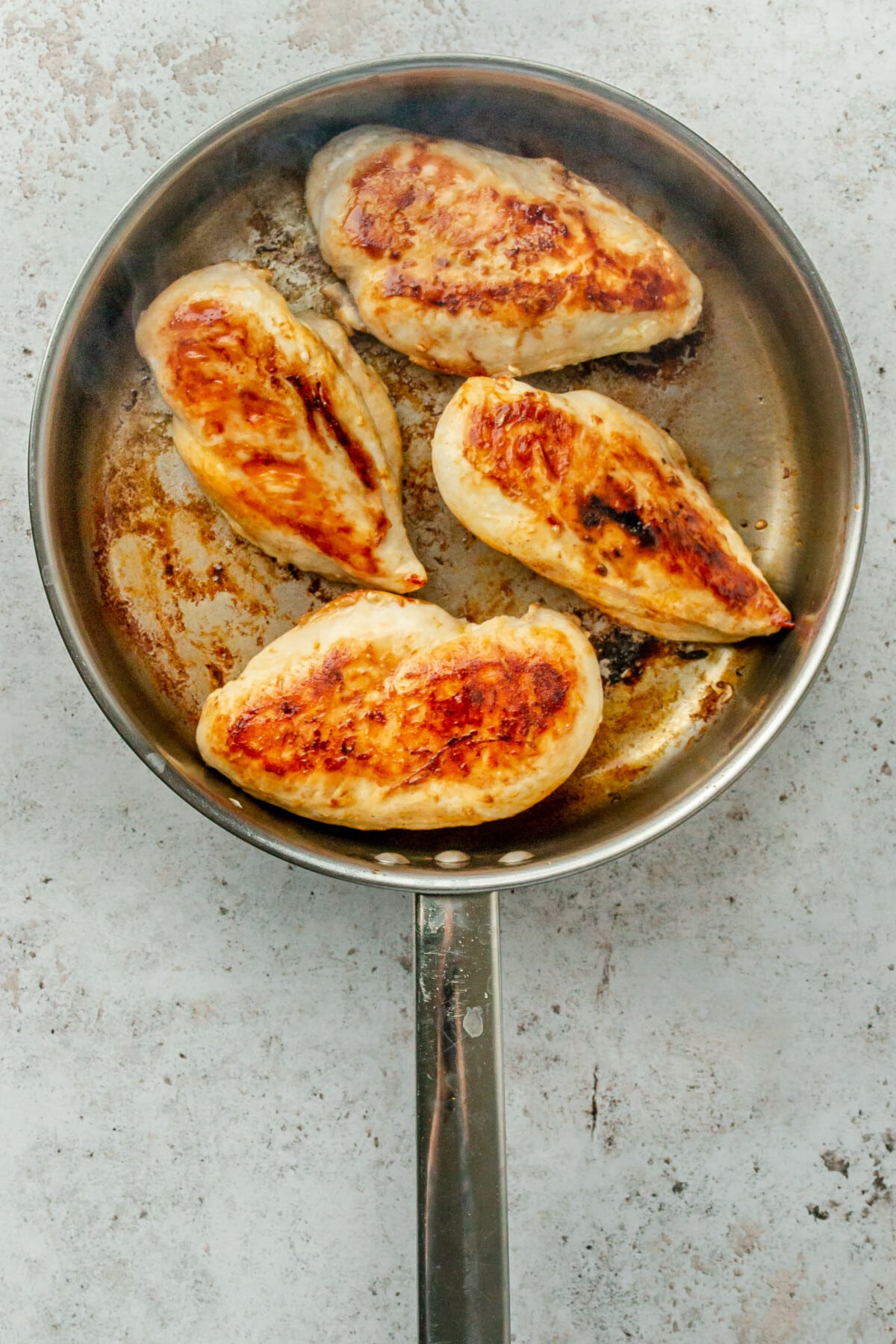 Brown cooked chicken breasts sit in a stainless steel frying pan on a light grey surface.