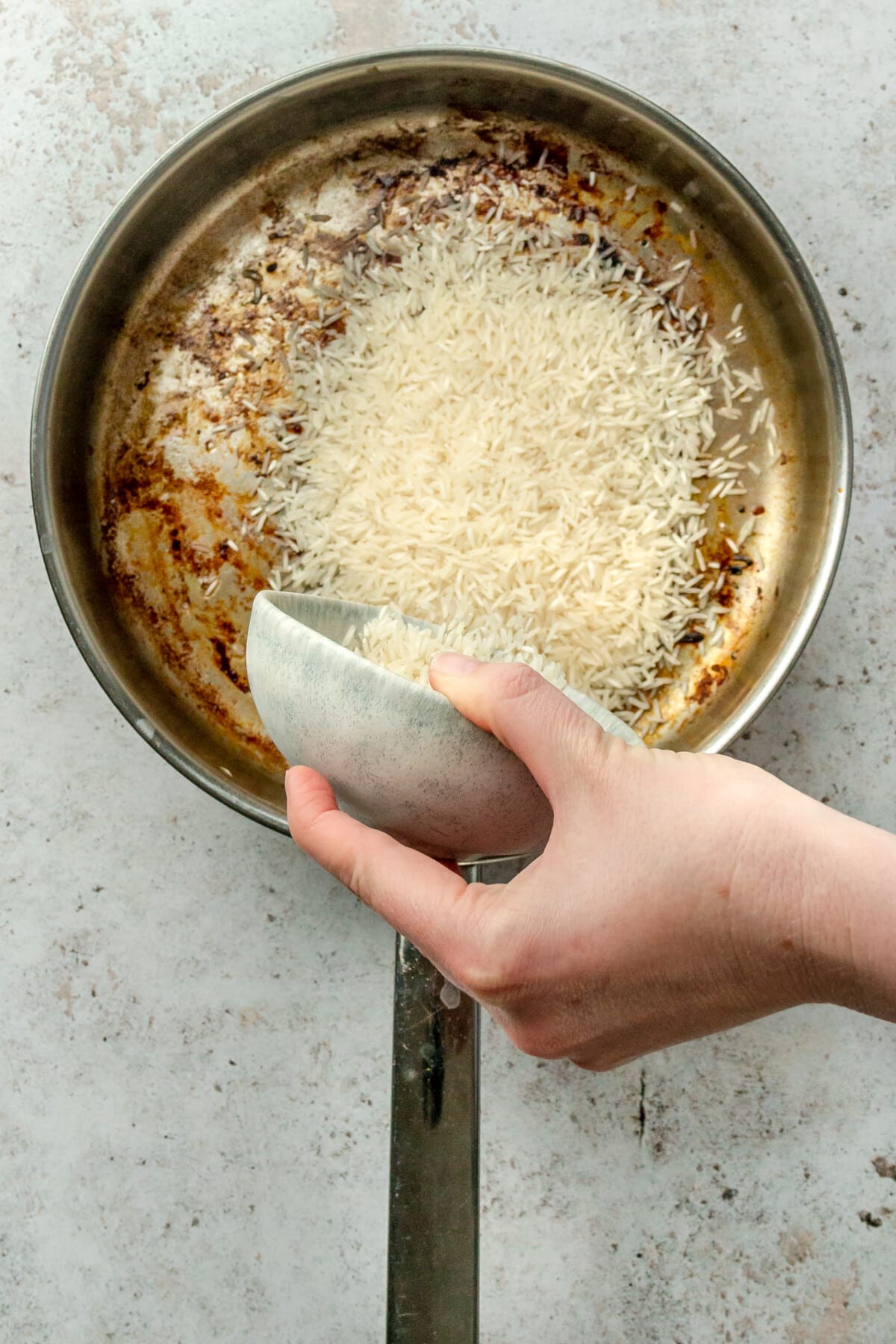 Rice is tossed into a stainless steel frying pan on a light grey surface.