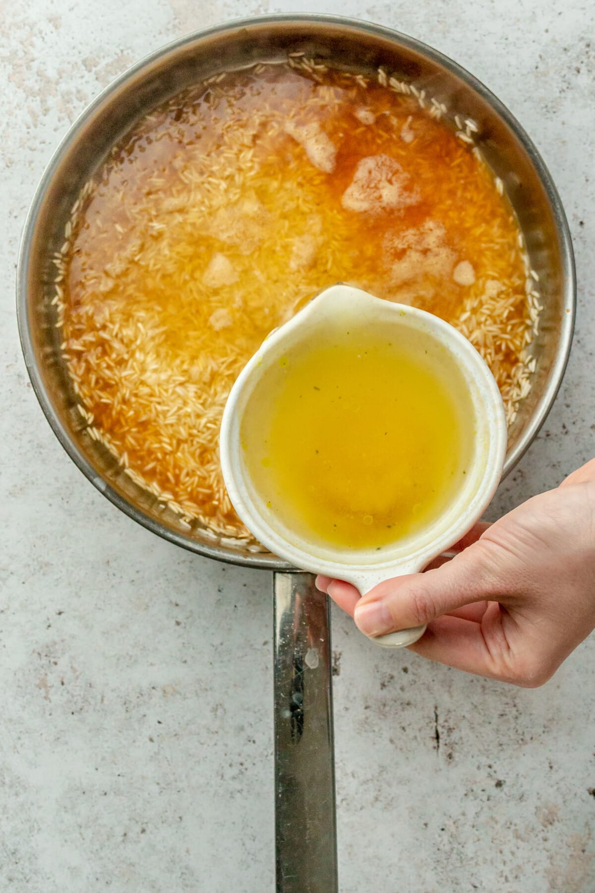 Chicken stock is poured over rice sitting in a stainless steel frying pan on a light grey surface.