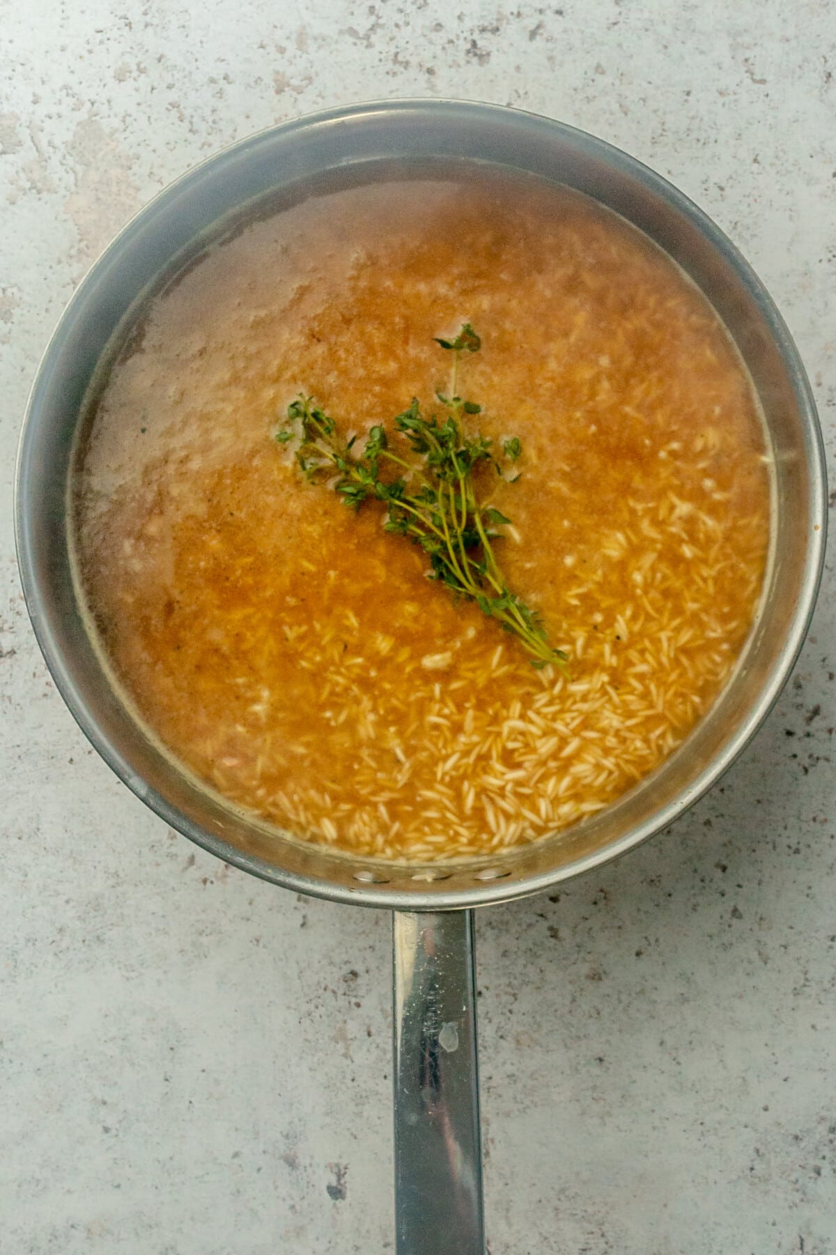 Thyme sprigs sit on top of rice and chicken stock in a stainless steel frying pan on a light grey surface.