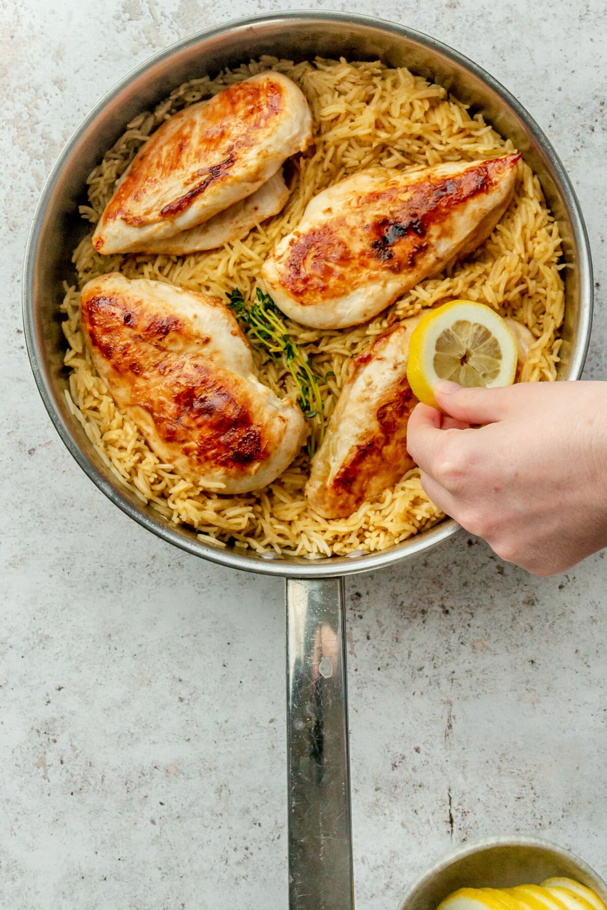 Slices of lemon are placed on top of cooked chicken breasts and rice in a stainless steel pan on a light grey surface.