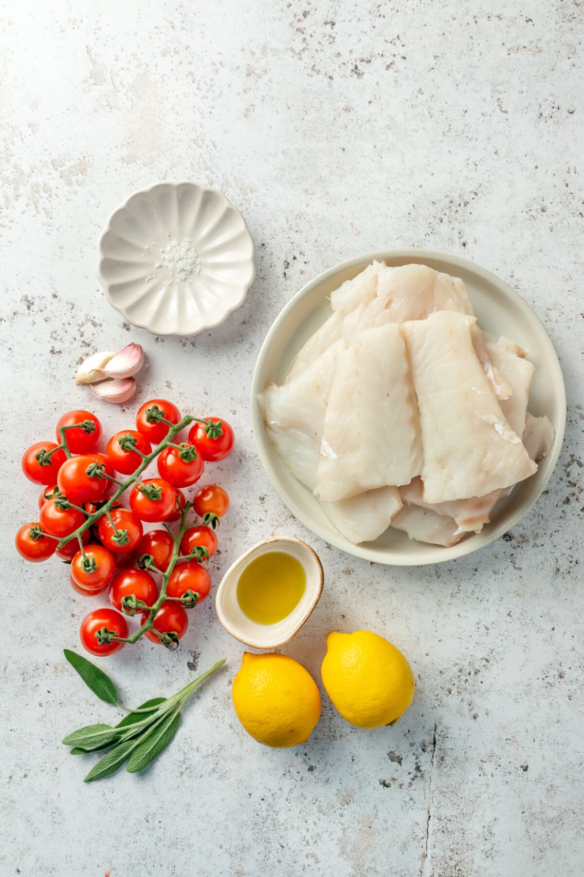 Ingredients for lemon garlic baked cod sit in a variety of bowls on a light grey surface.