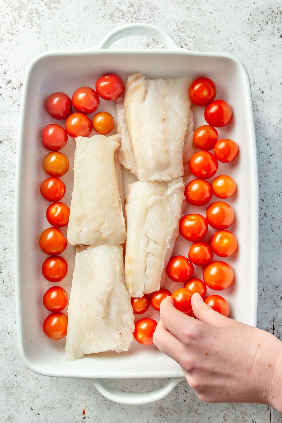 Cherry tomatoes are shown being added to a white rectangular baking dish which holds four cod filets.