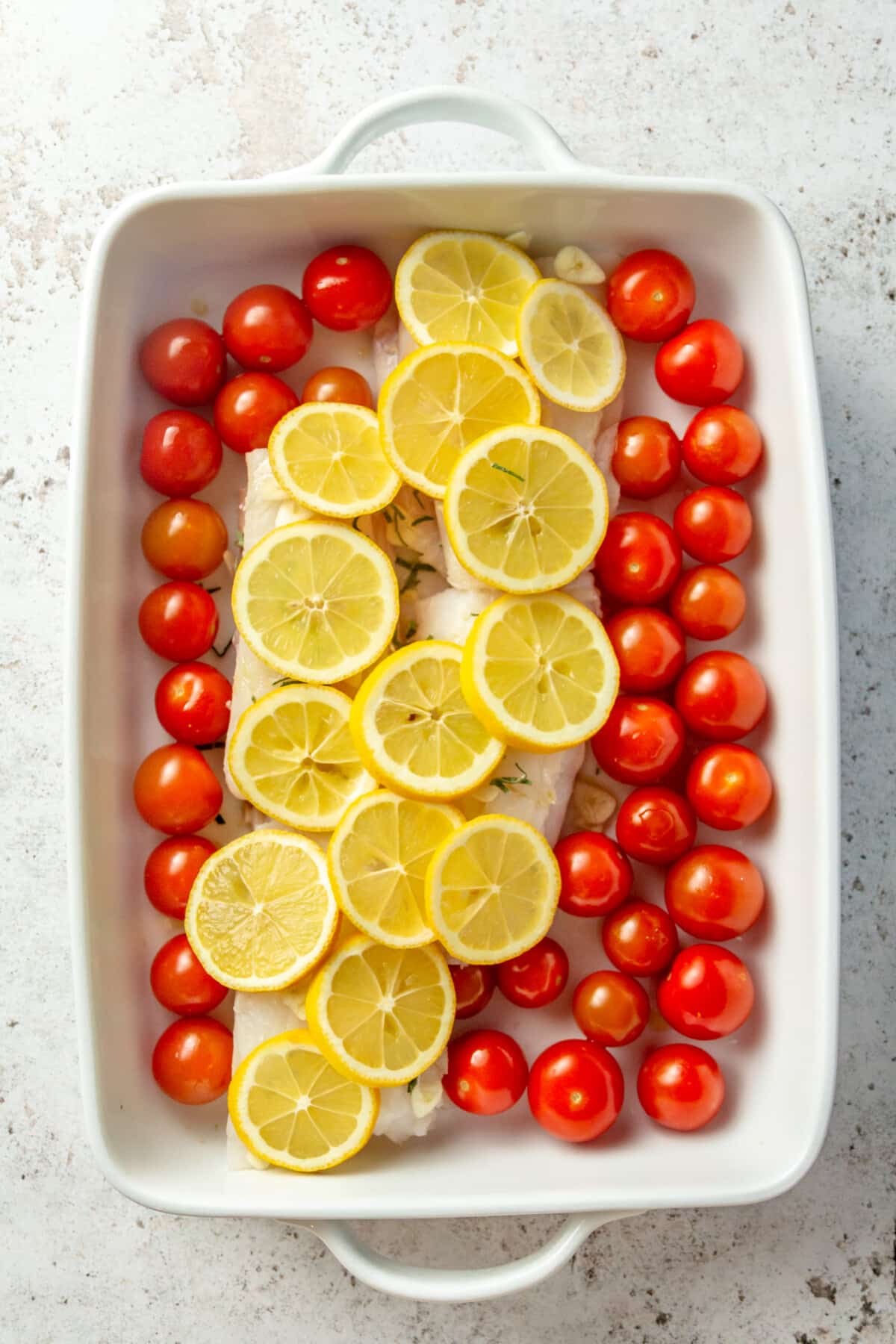 Cod filets and cherry tomatoes sit in a white rectangular baking dish. Slices of lemon sit on top.
