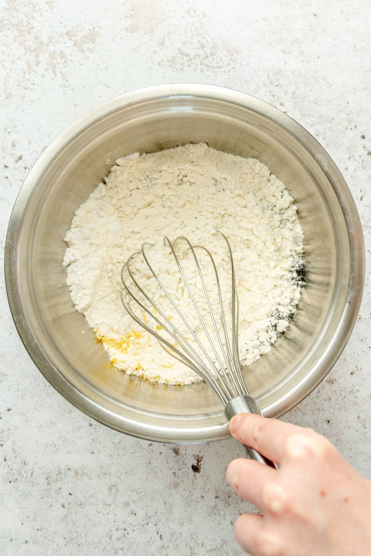 Dry ingredients are whisked for a lemon poppy seed loaf in a stainless steel bowl on a light grey surface.
