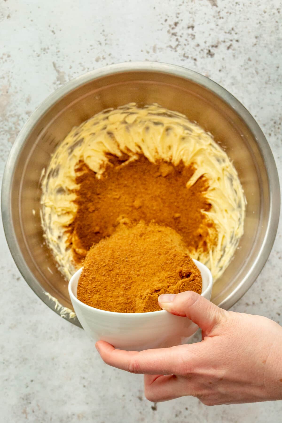 Coconut sugar is added to whipped butter in a stainless steel bowl on a light grey surface.