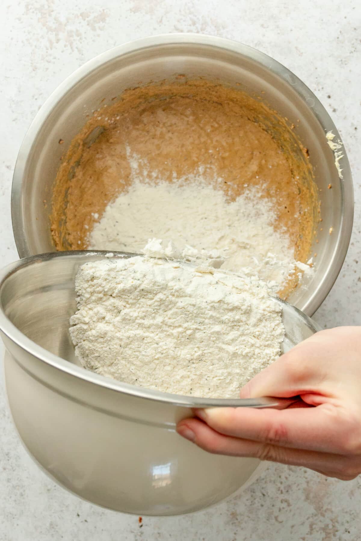 A mixture of dry ingredients are added to a whipped butter, sugar and egg mixture in a stainless steel bowl on a light grey surface.