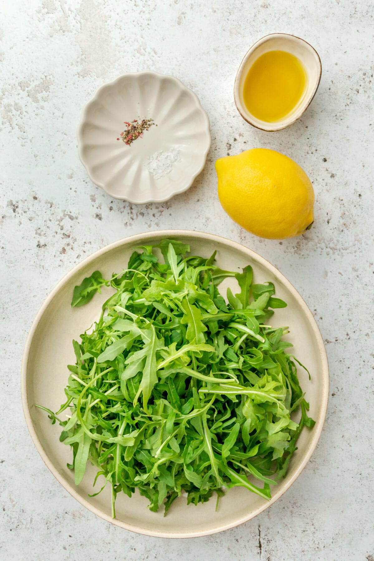 Ingredients for arugula salad sit in a variety of plates on a light grey surface.