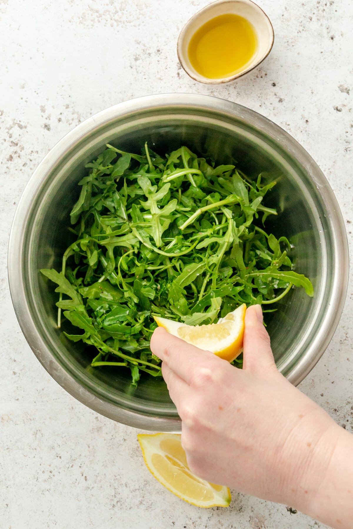 A lemon wedge is squeezed over an arugula salad in a stainless steel bowl on a light grey surface.