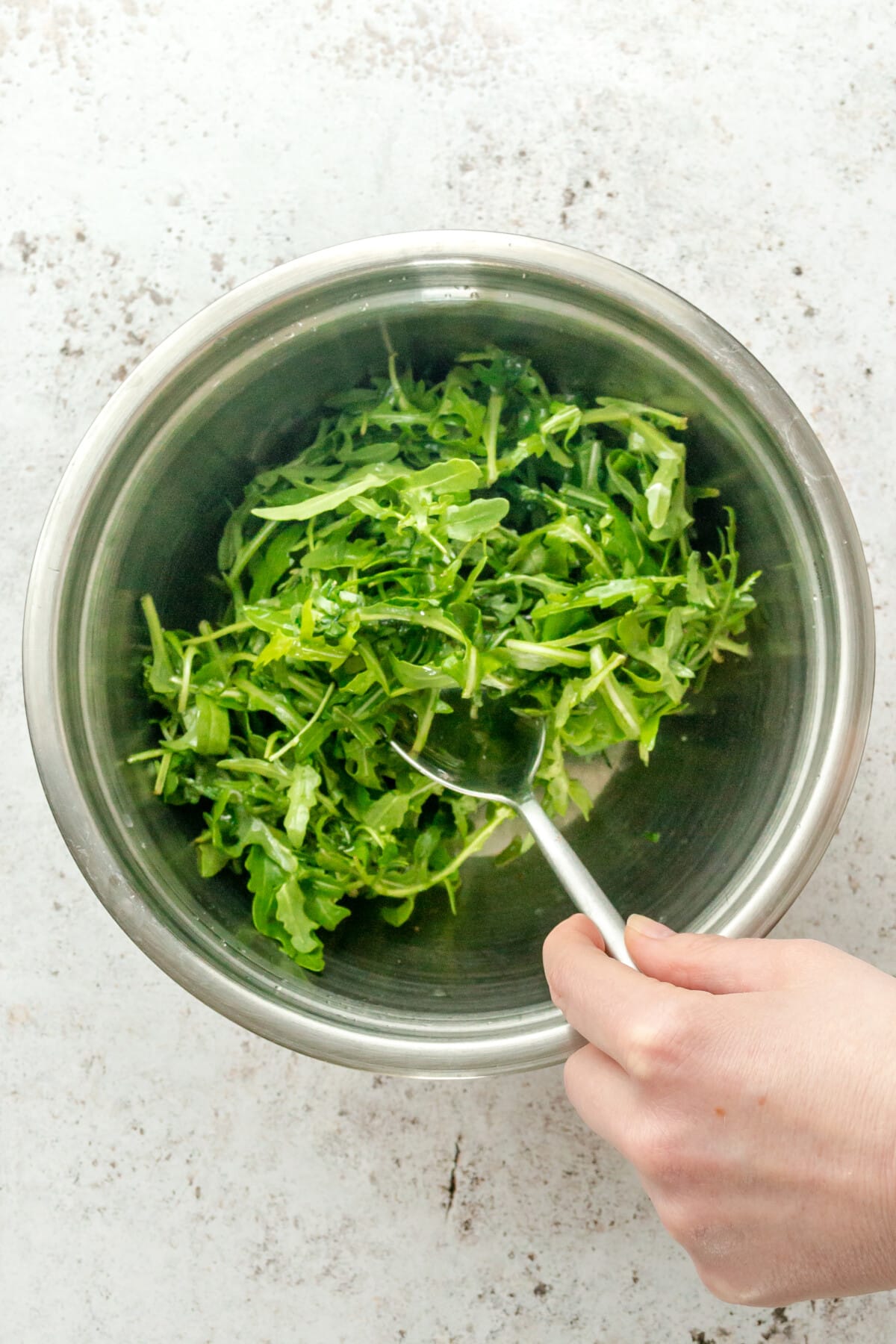 Arugula is stirred with a light dressing sitting in a stainless steel bowl on a light grey surface.