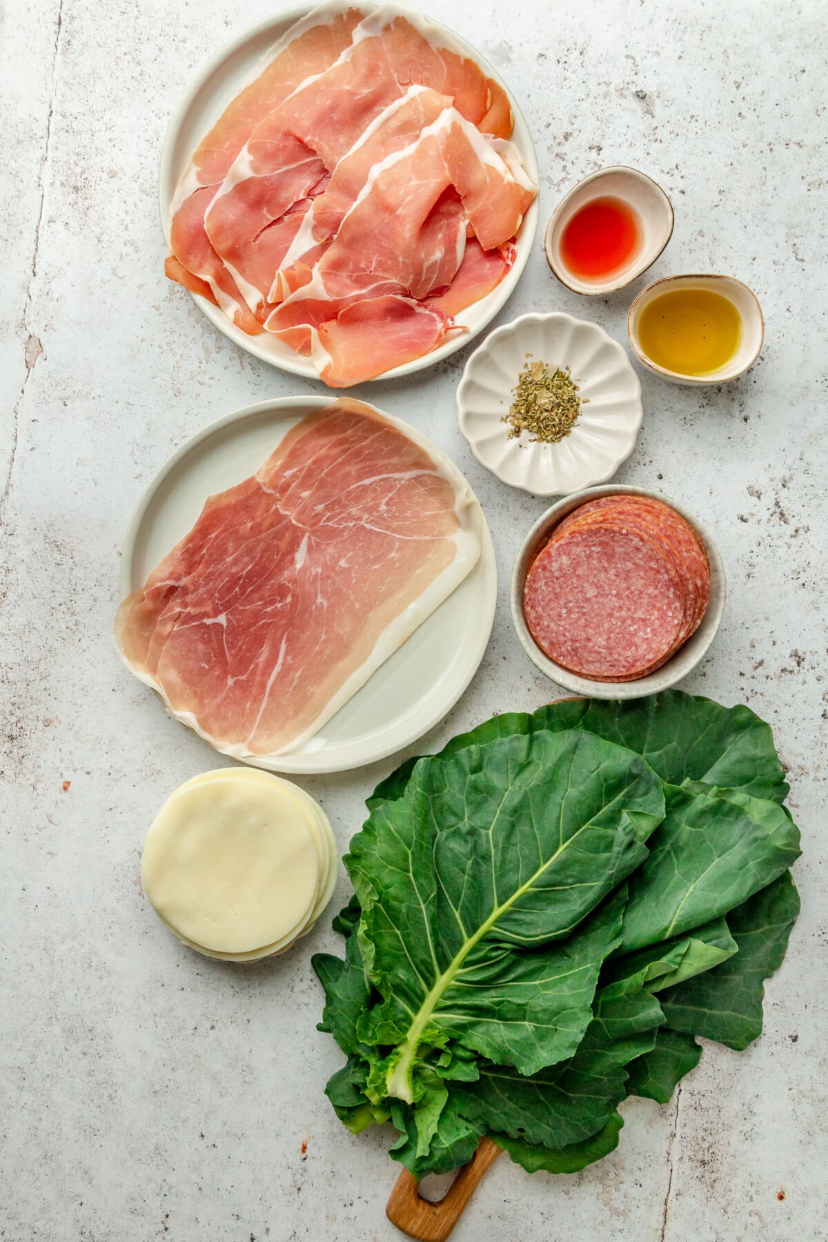 Ingredients for make ahead Italian collard wraps sit in a variety of plates and bowls on a light grey surface.