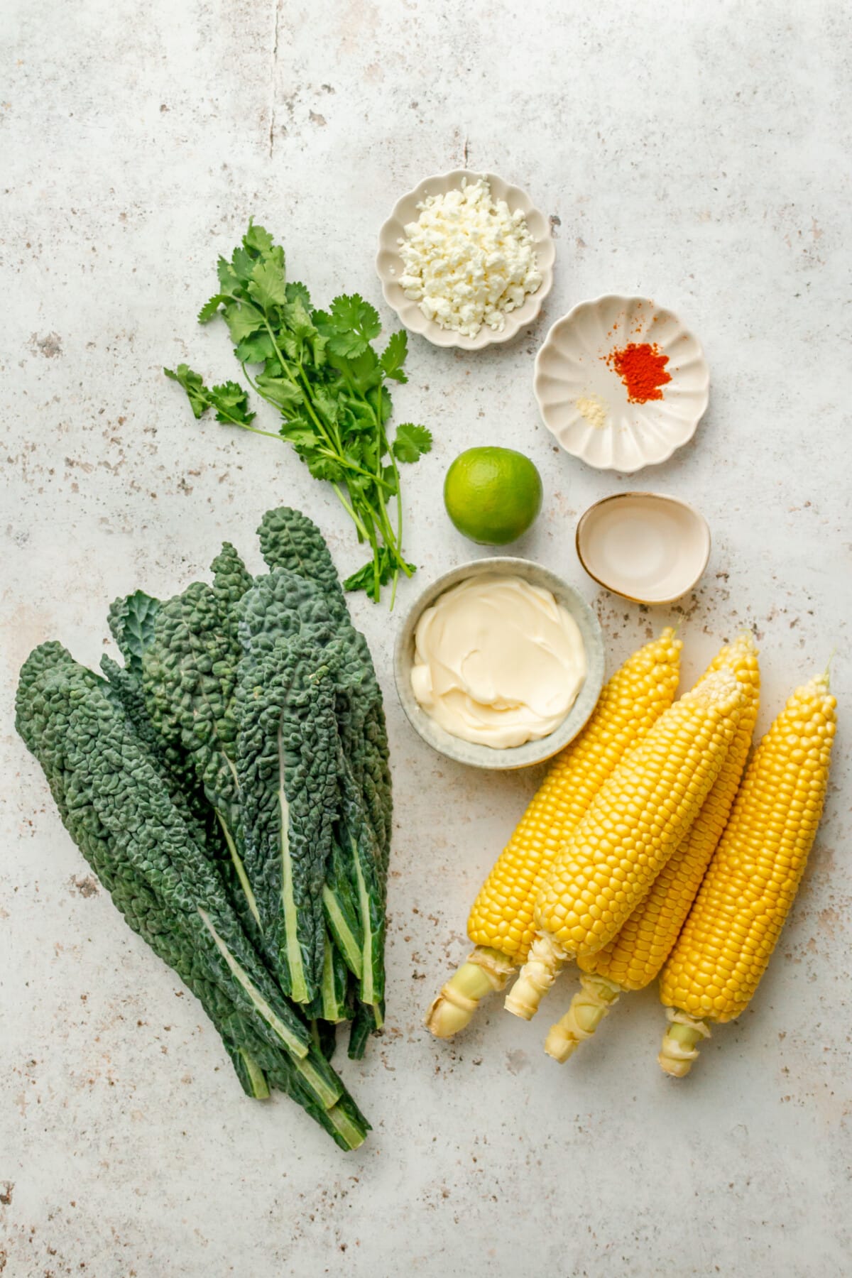 Ingredients for Mexican street corn and kale slaw sit in a variety of bowls on a light grey surface.