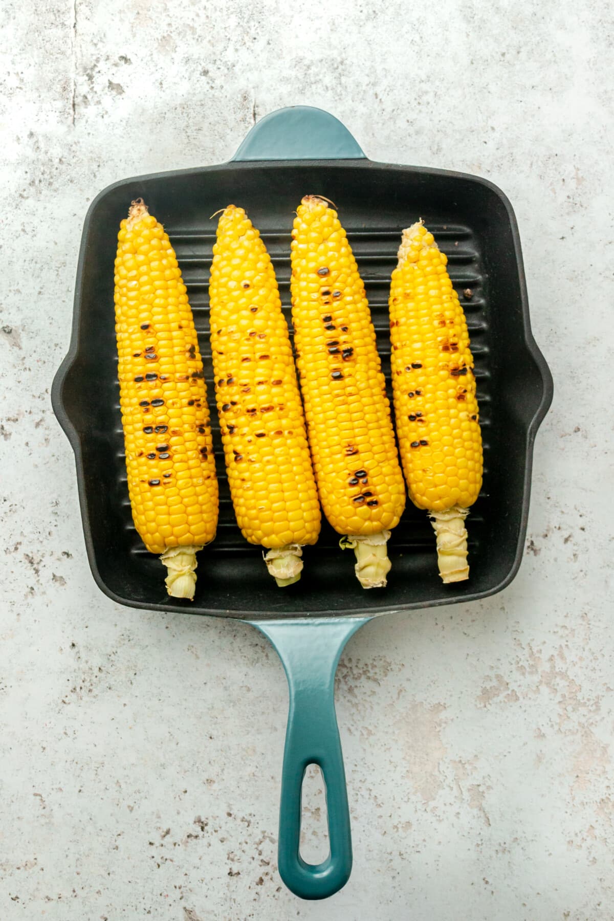 Grilled corn sits in a grill skillet on a light grey surface.