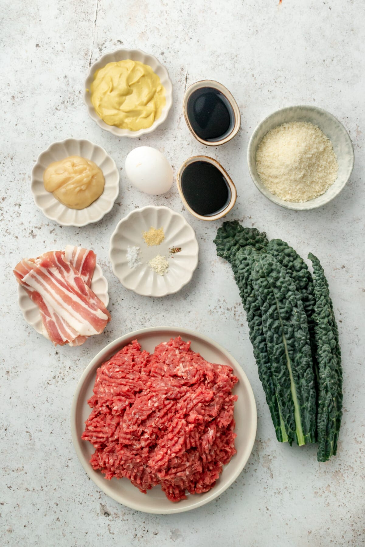 Ingredients for mini meatloaves with balsamic glaze are laid out in a variety of small plates and bowls on a light grey colored surface.