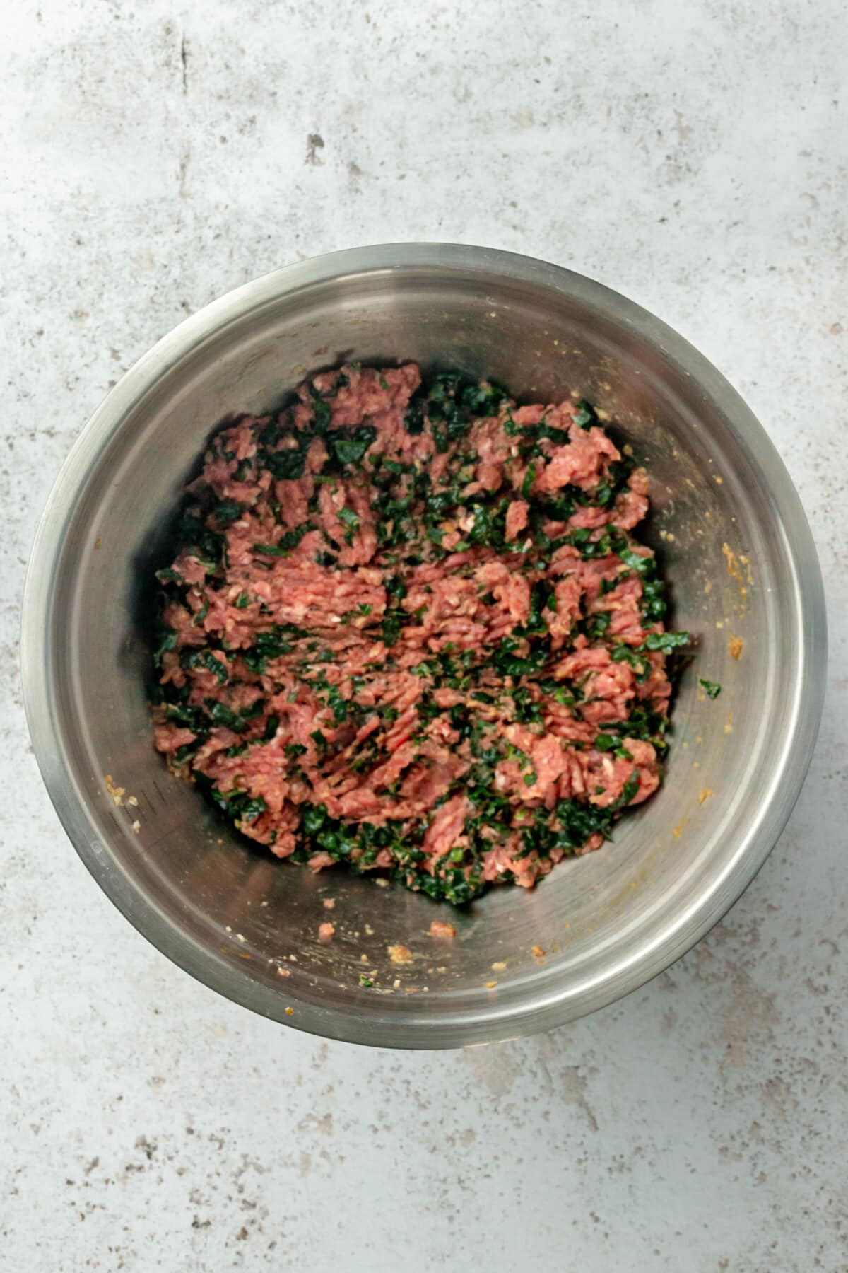 A meatloaf mixture with shredded kale sits in a stainless steel bowl on a light grey colored surface.