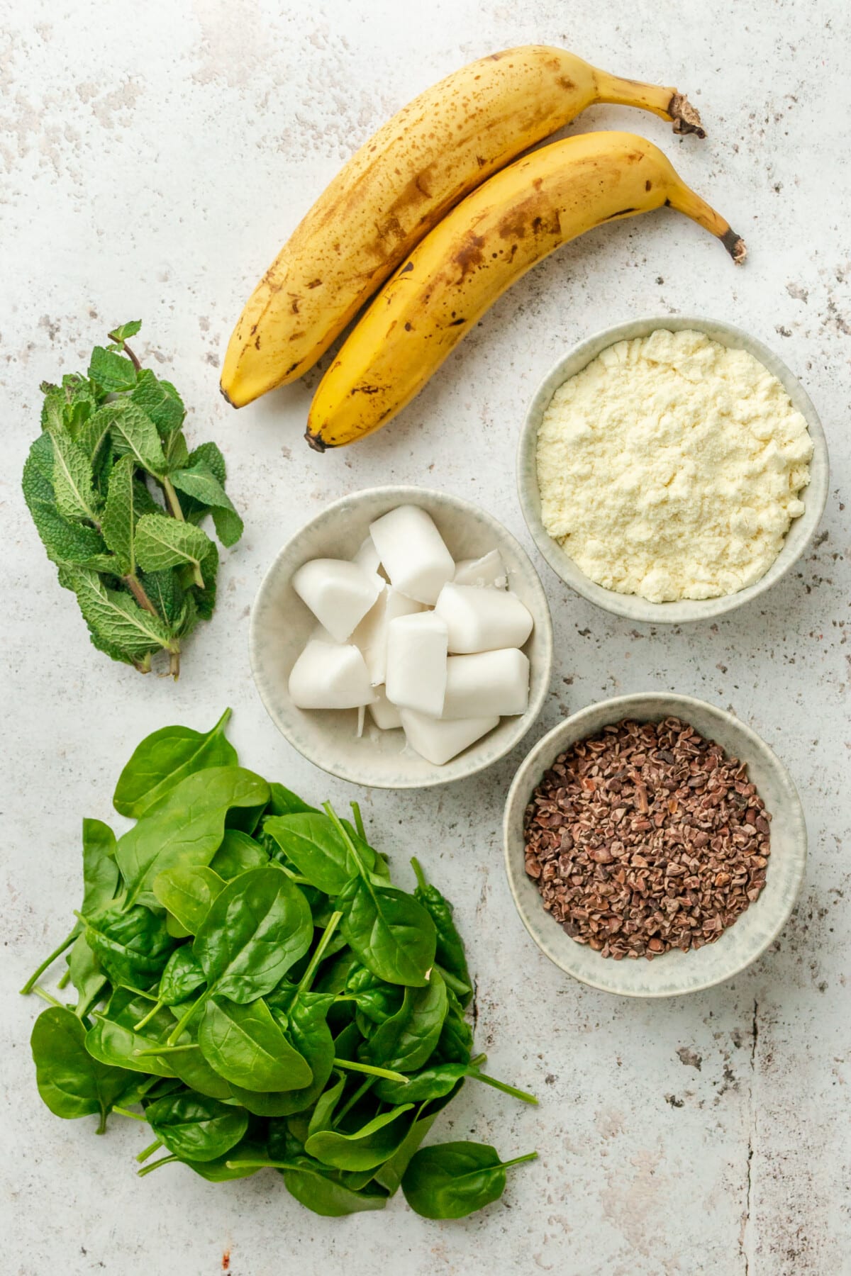 Ingredients for mint chocolate chip green smoothies are laid out in a variety of small plates on a light grey colored surface.
