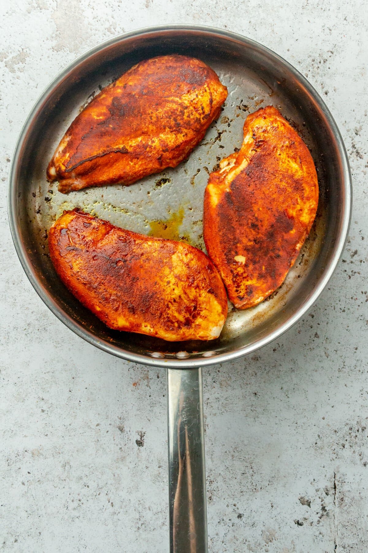 Cooked spiced chicken breasts sit in a stainless steel frying pan on a light grey surface.