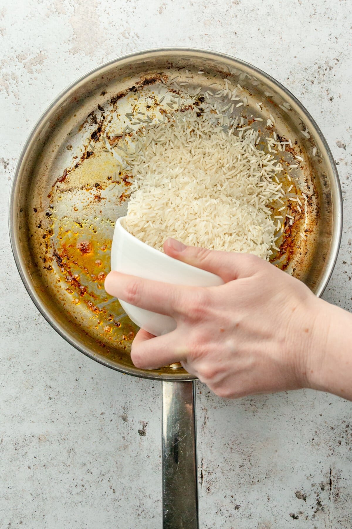 Rice is tossed into a stainless steel frying pan on a light grey surface.