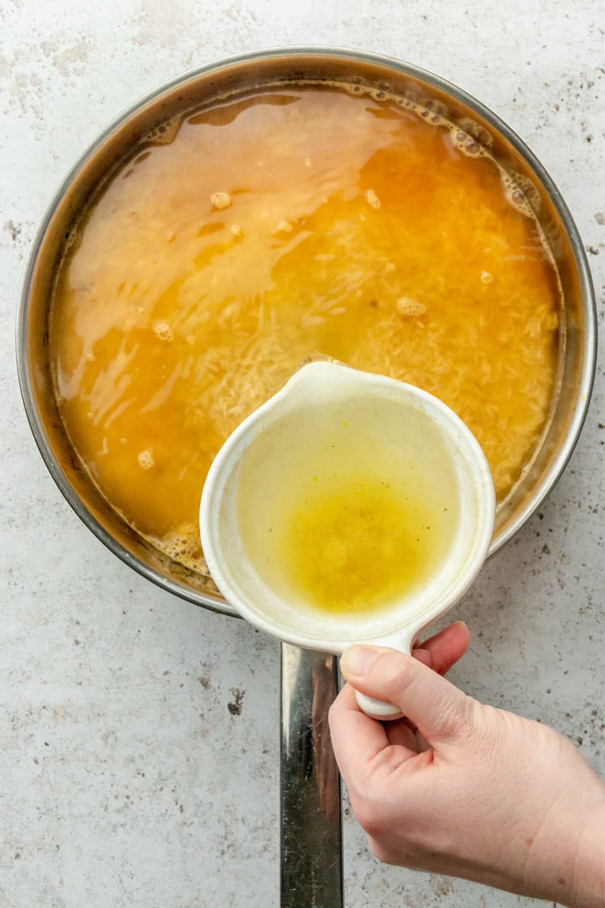 Chicken stock is poured into a stainless steel frying pan on a light grey surface.