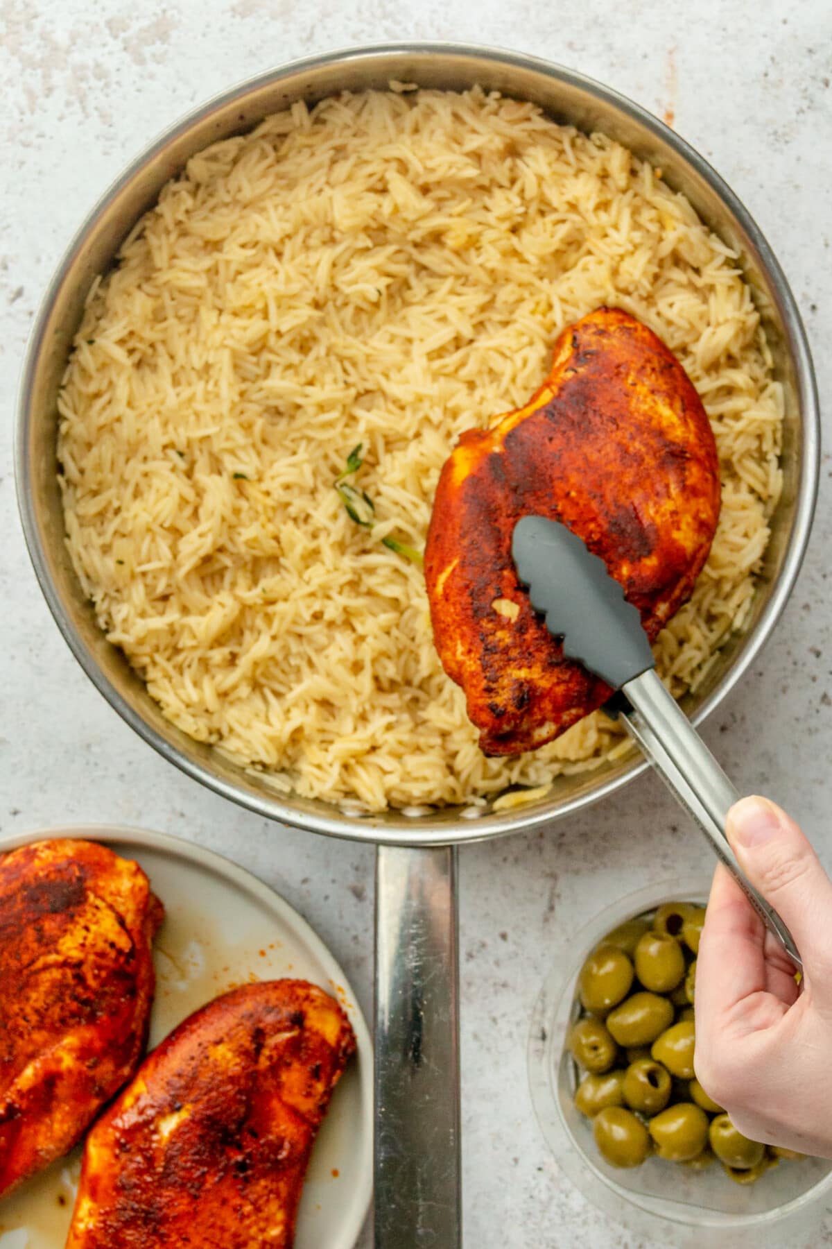 Spiced chicken is placed on top of rice in a stainless steel frying pan on a light grey surface.